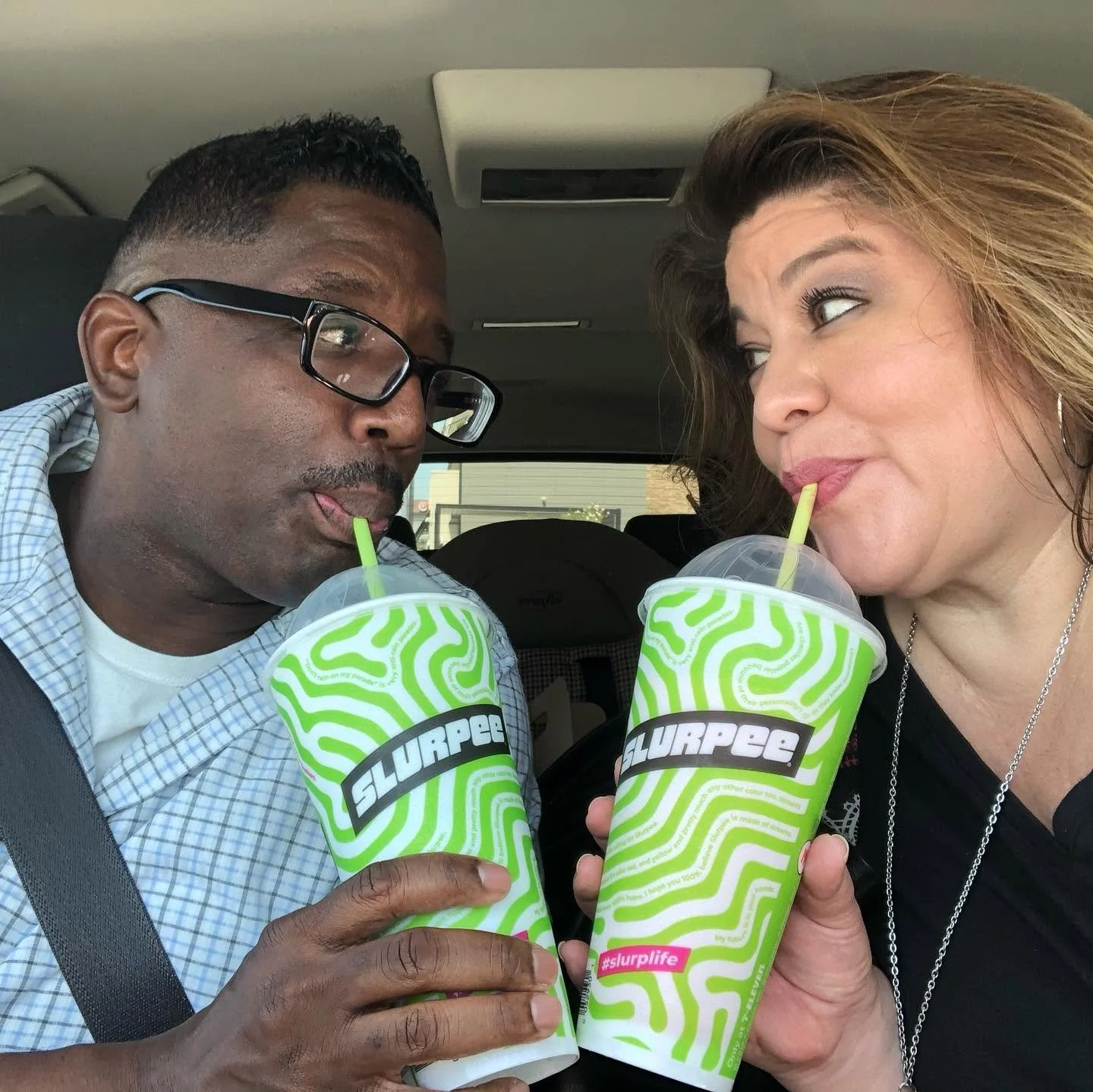Two people sitting in a car sharing slurpees with green and white swirl packaging, looking at each other and drinking through straws.