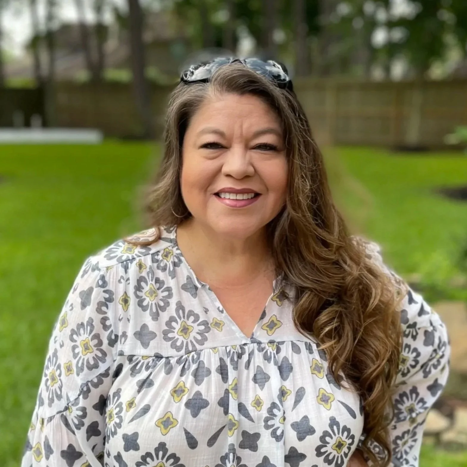 A woman with long curly hair wearing a patterned blouse, smiling outdoors in a green backyard with a wooden fence in the background.