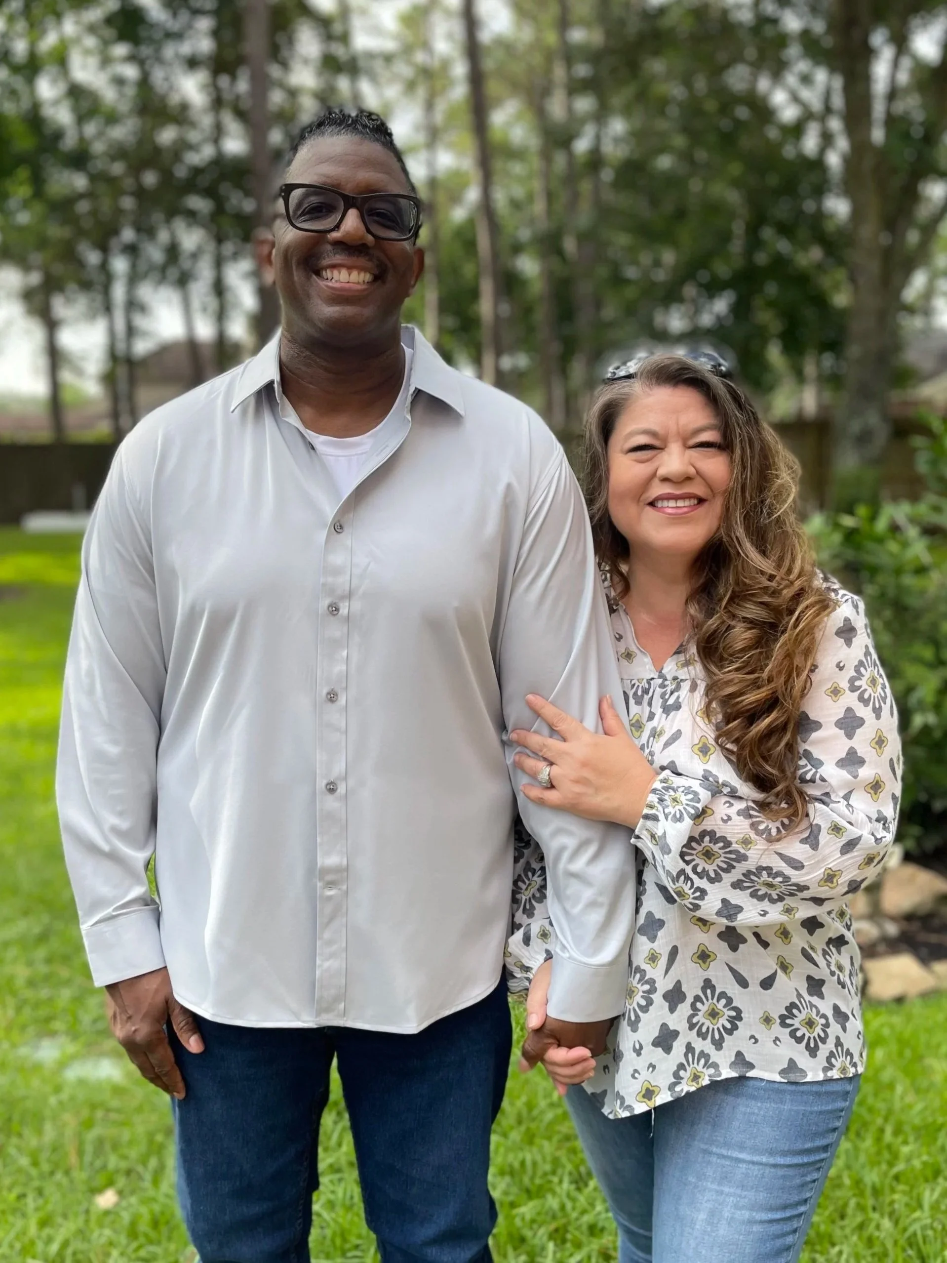 A smiling Black man wearing glasses and a white button-up shirt standing outdoors with a smiling white woman with long curly hair and glasses, who is holding his hand. They are in a grassy area with trees in the background.