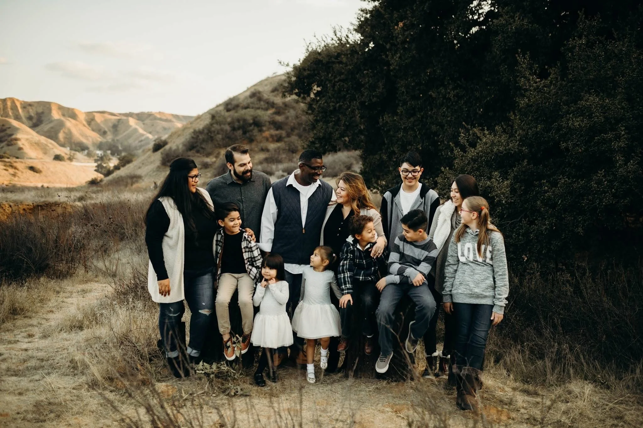 A group of diverse people including children and adults standing outdoors in a natural landscape with grassy fields, trees, and rolling hills under a partly cloudy sky, smiling and enjoying each other's company.