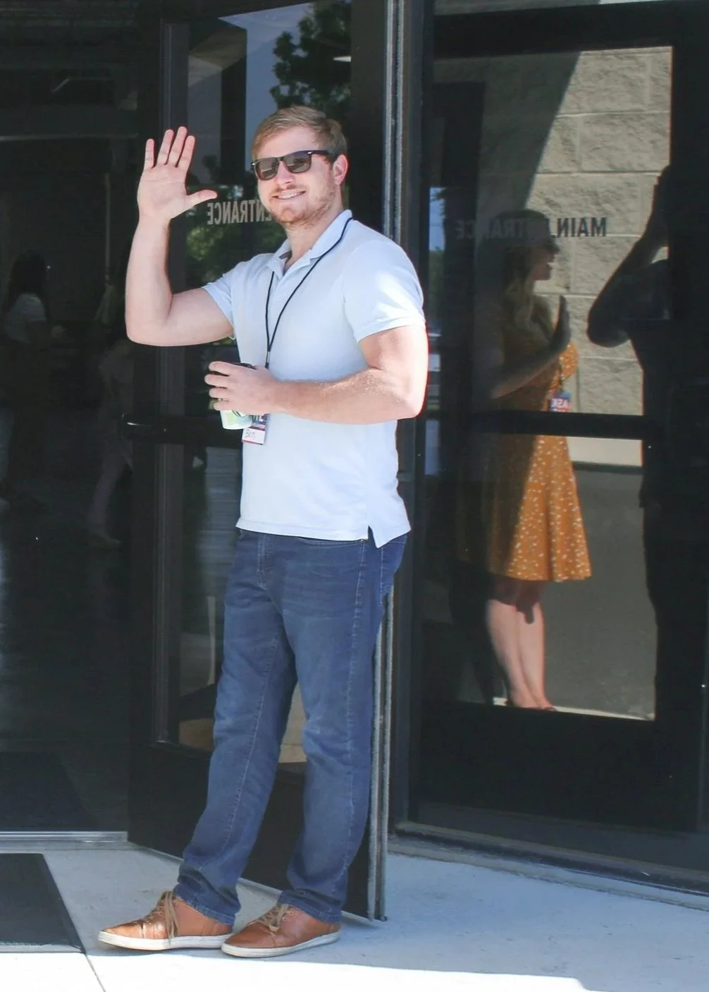A man in sunglasses, a white polo shirt, and jeans waving and smiling outside a building with glass doors. Reflections of a woman in a yellow dress and other people are visible in the glass.