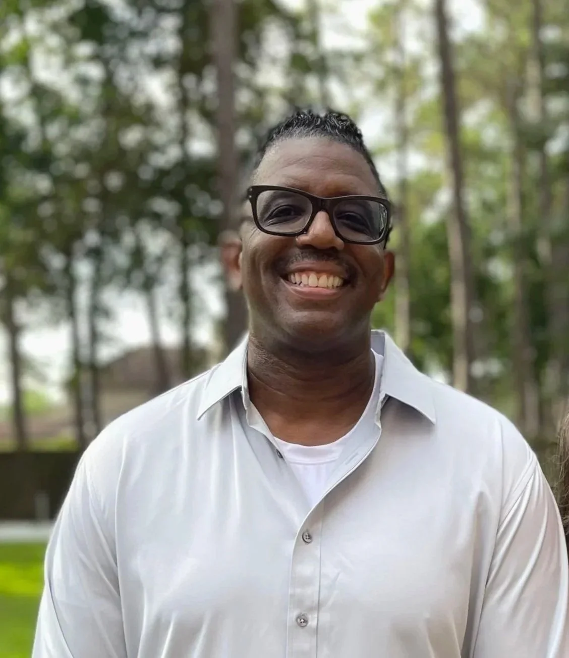 Smiling man with glasses wearing a white button-up shirt outdoors with trees in the background.