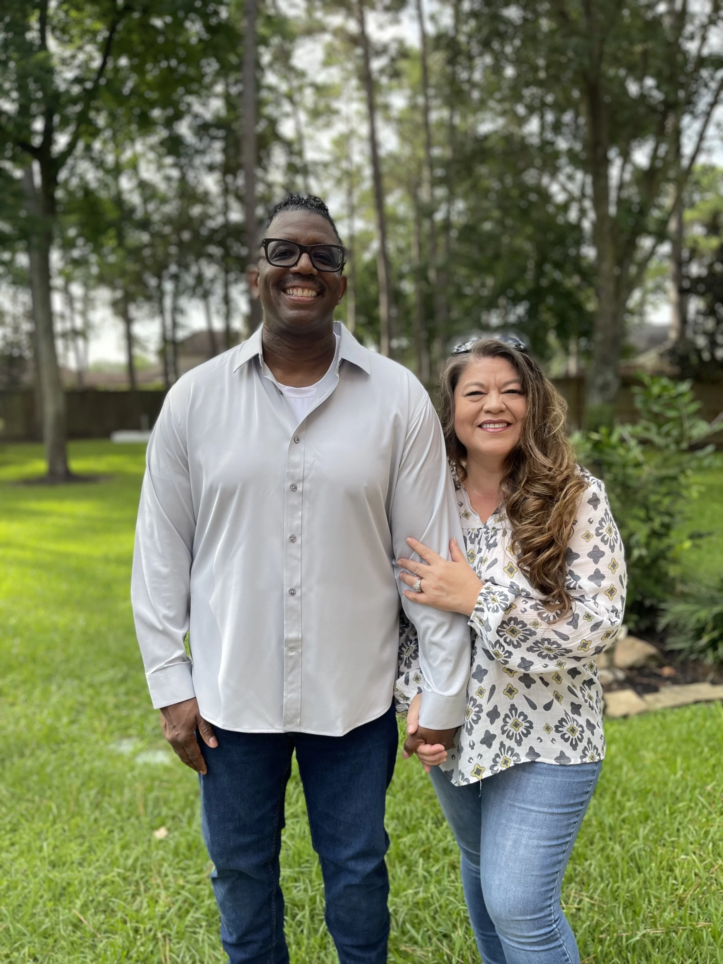 A smiling man and woman standing outdoors in a park-like area with green grass and tall trees background. The man is wearing glasses, a white button-up shirt, and blue jeans. The woman has long curly hair, is wearing a patterned blouse and blue jeans, and is holding the man's arm.