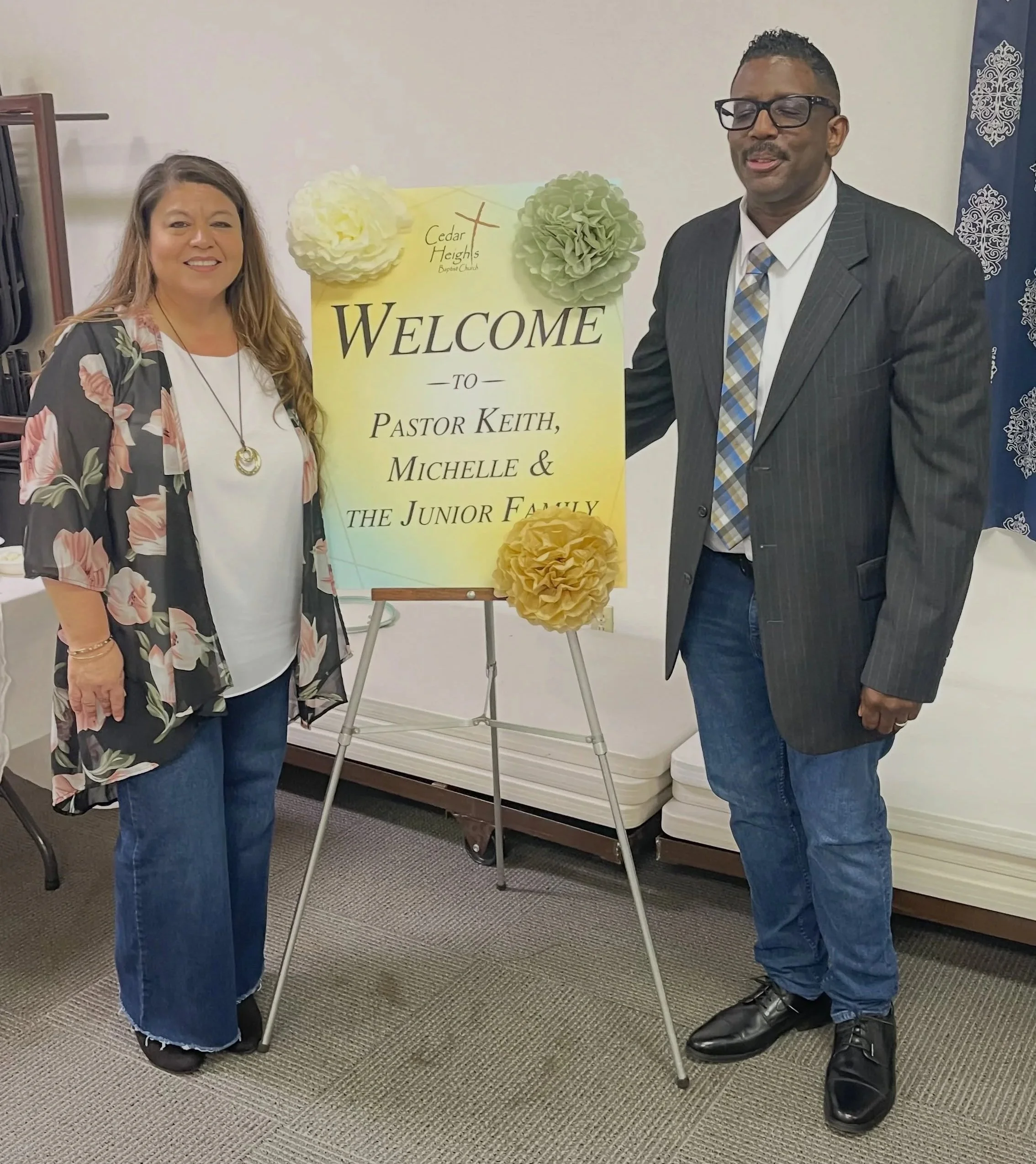 Two people, a woman and a man, are standing on either side of a welcome sign for Cedar Heights Baptist Church. The woman has long hair, wears glasses, a floral kimono, and a white shirt with a necklace. The man has short curly hair, wears glasses, a striped suit jacket, white shirt, striped tie, and jeans. They are smiling and holding a piece of the sign, which is decorated with large paper flowers and a yellow tissue paper pom-pom at the bottom.