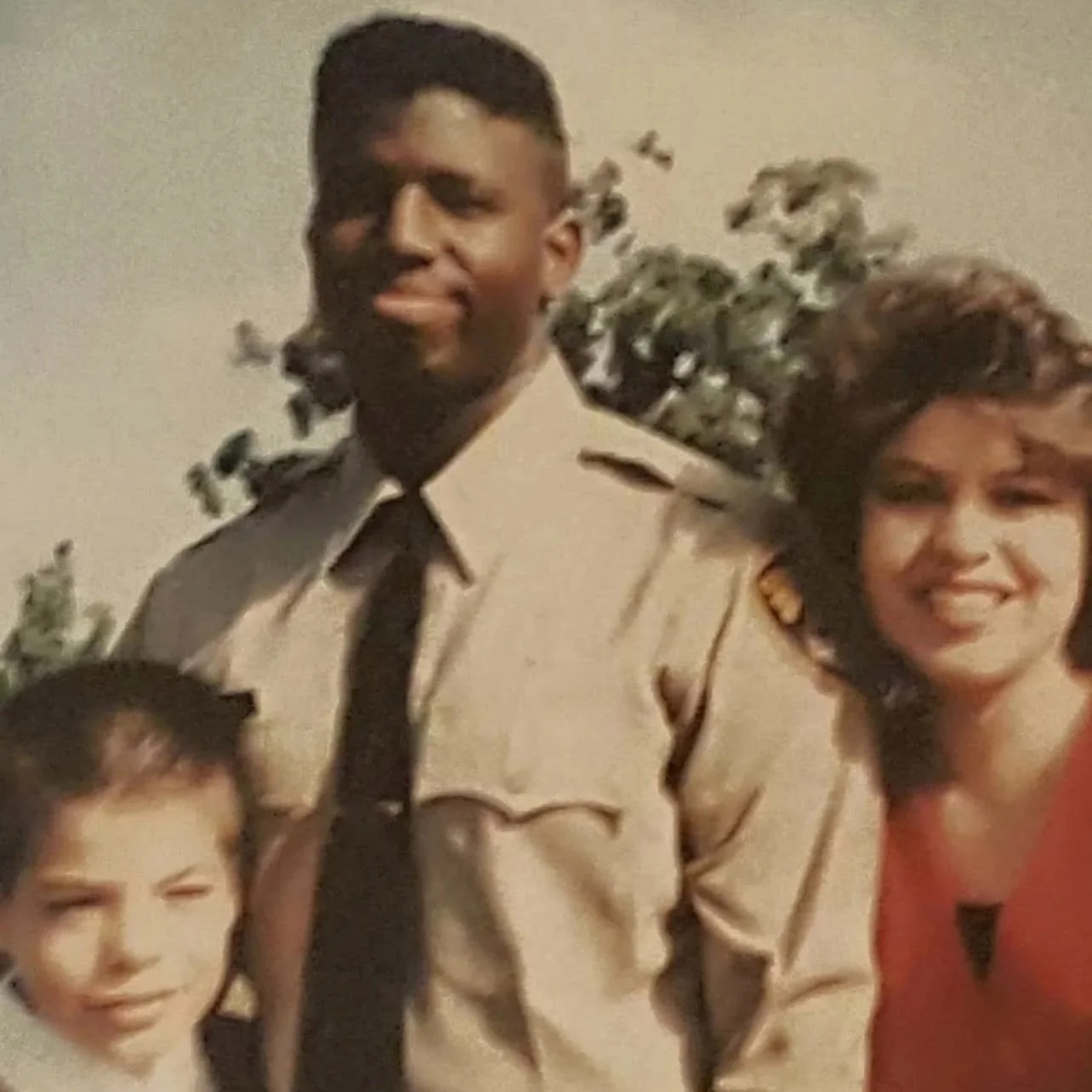 A group of people, including a young boy, a young man in a uniform, and a woman, smiling at a photo. The young man is dressed in a beige uniform with a tie, and the woman has curly hair and is wearing a red top. The background has some greenery and a plain wall.
