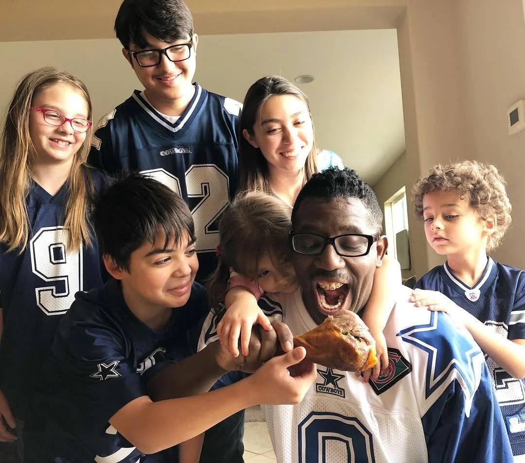 A group of children and a man wearing Dallas Cowboys football jerseys gathered around as the man is about to eat a large piece of food, with the children helping him hold it.