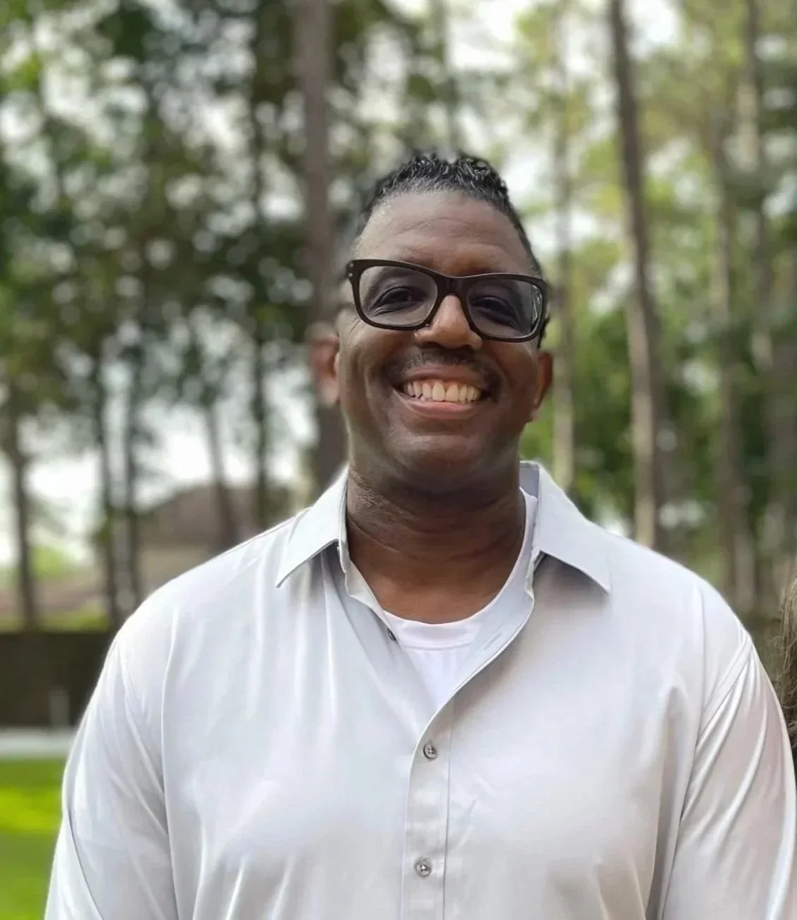 Smiling man with glasses wearing a white button-up shirt outdoors with trees in the background.