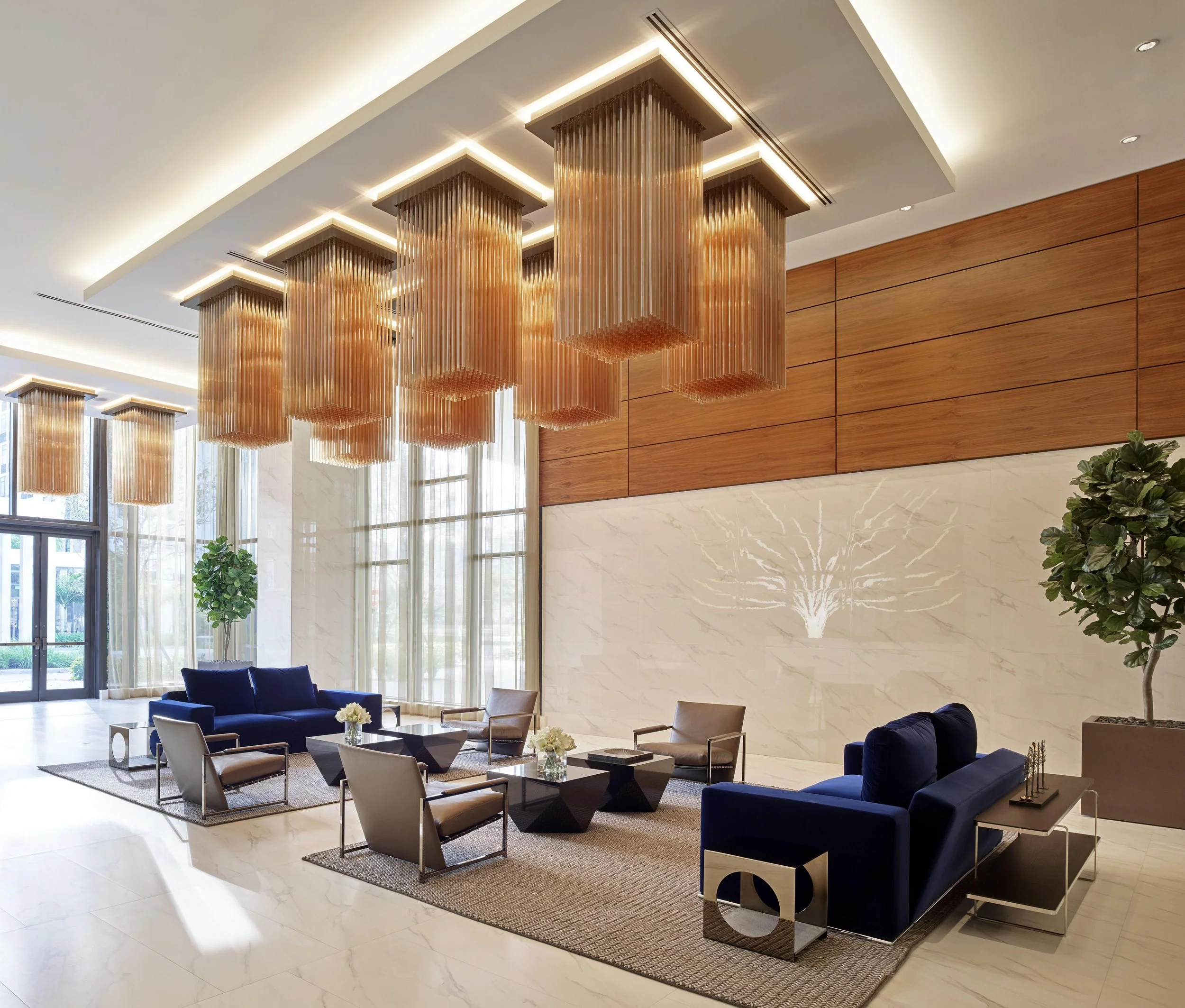 The image shows a modern hotel lobby or lounge area with high ceilings featuring decorative hanging light fixtures and large windows with sheer curtains. There are several seating arrangements with sofas, armchairs, and coffee tables, and a wooden accent wall on the right side. The space is bright and stylish with a polished marble floor.