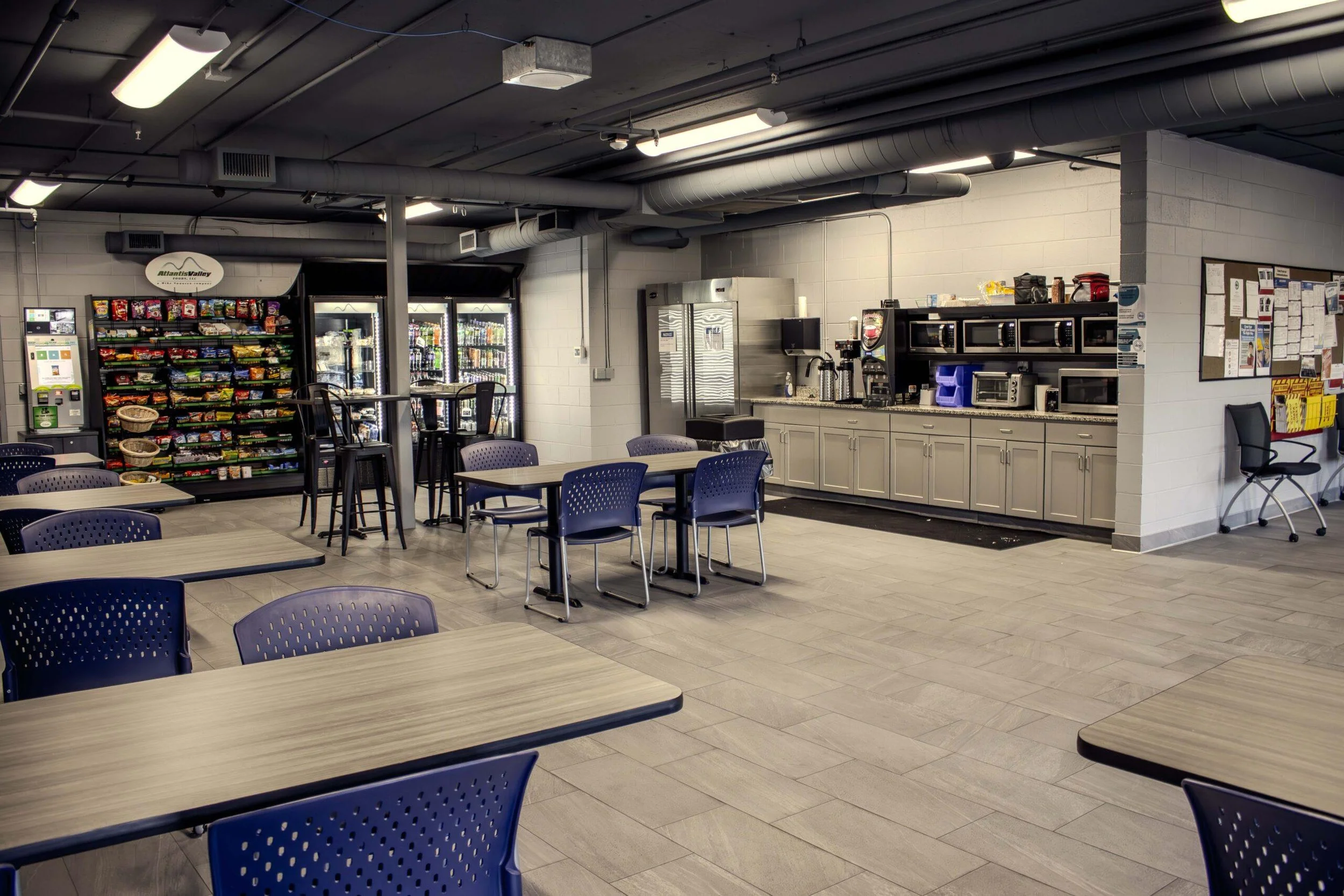 Indoor break room or lounge area with tables and chairs, refrigerator, vending machines, microwave, and bulletin board, with industrial ceiling vents and lighting.