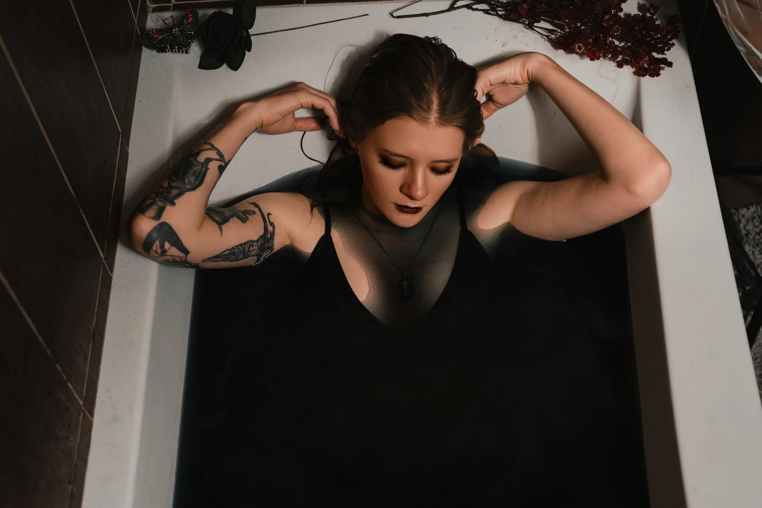 Woman with dark hair and tattoos relaxing in a bathtub filled with water, with her eyes closed and arms resting on her head, surrounded by decorative flowers and a black butterfly ornament.