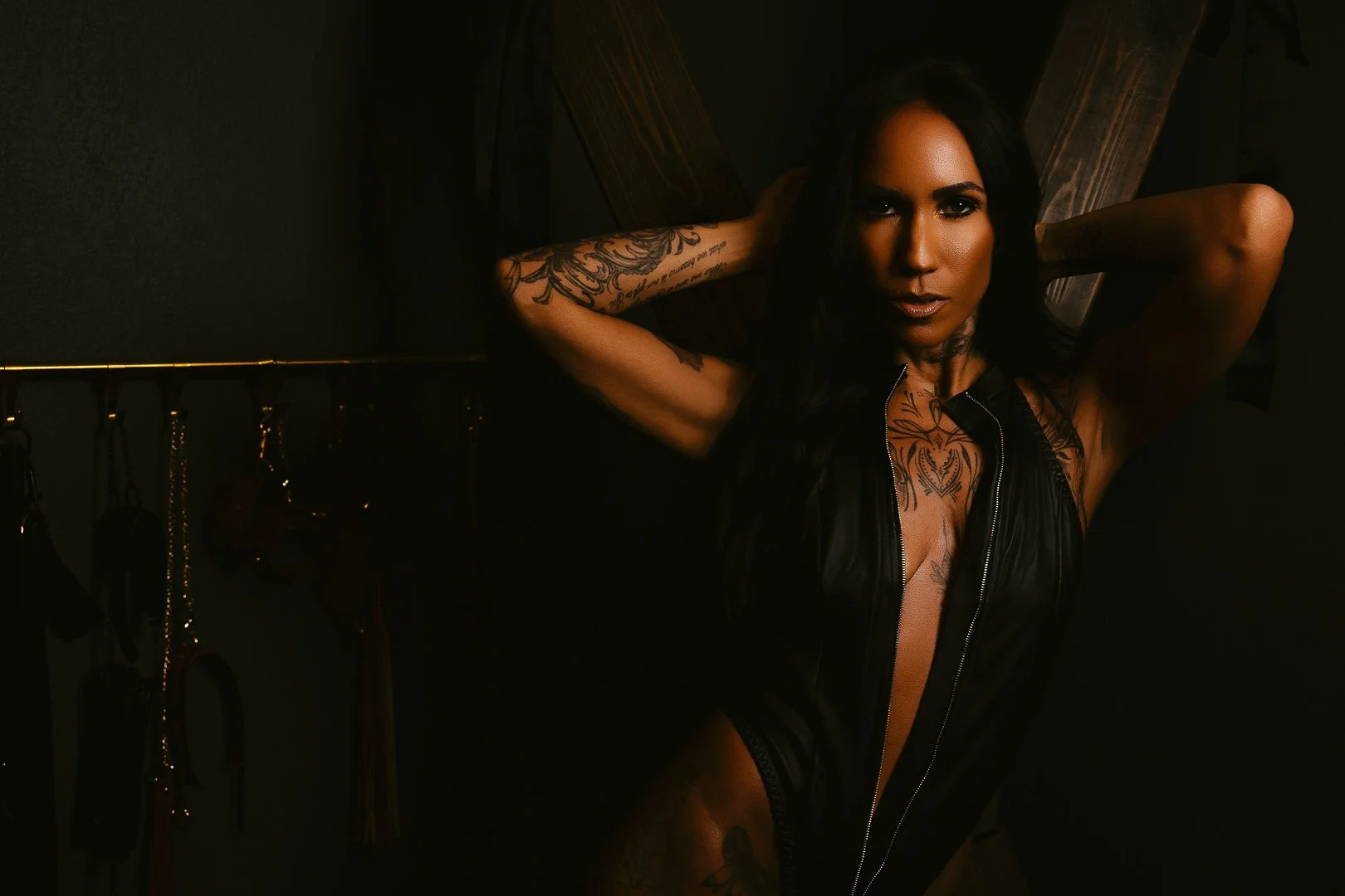A woman with dark hair and tattoos wearing a black leather outfit, posing with her hands behind her head in a dimly lit room.