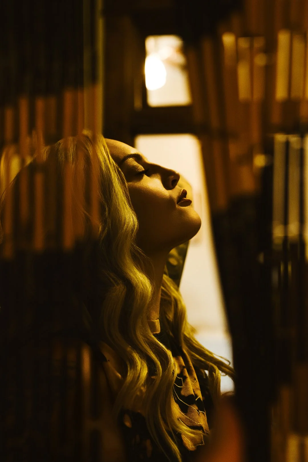 A woman with long, wavy blonde hair leaning against a bookshelf with her eyes closed, inside a dimly lit library or bookstore.