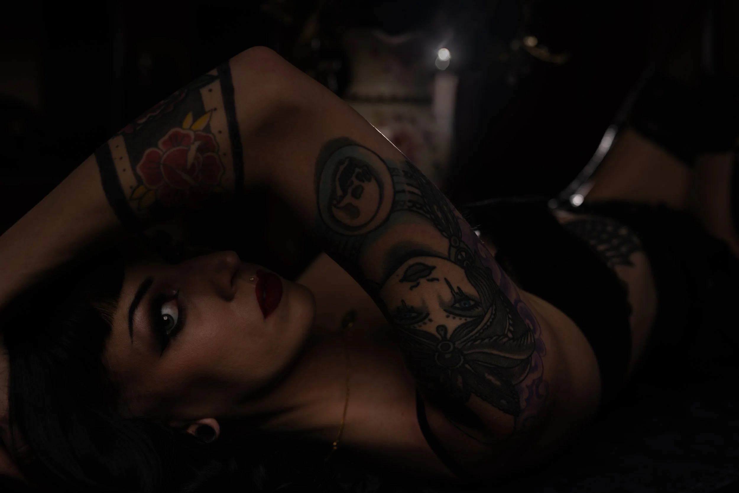 A woman with dark hair and tattoos lies on her side, looking at the camera with her head resting on her arm in a dark setting.