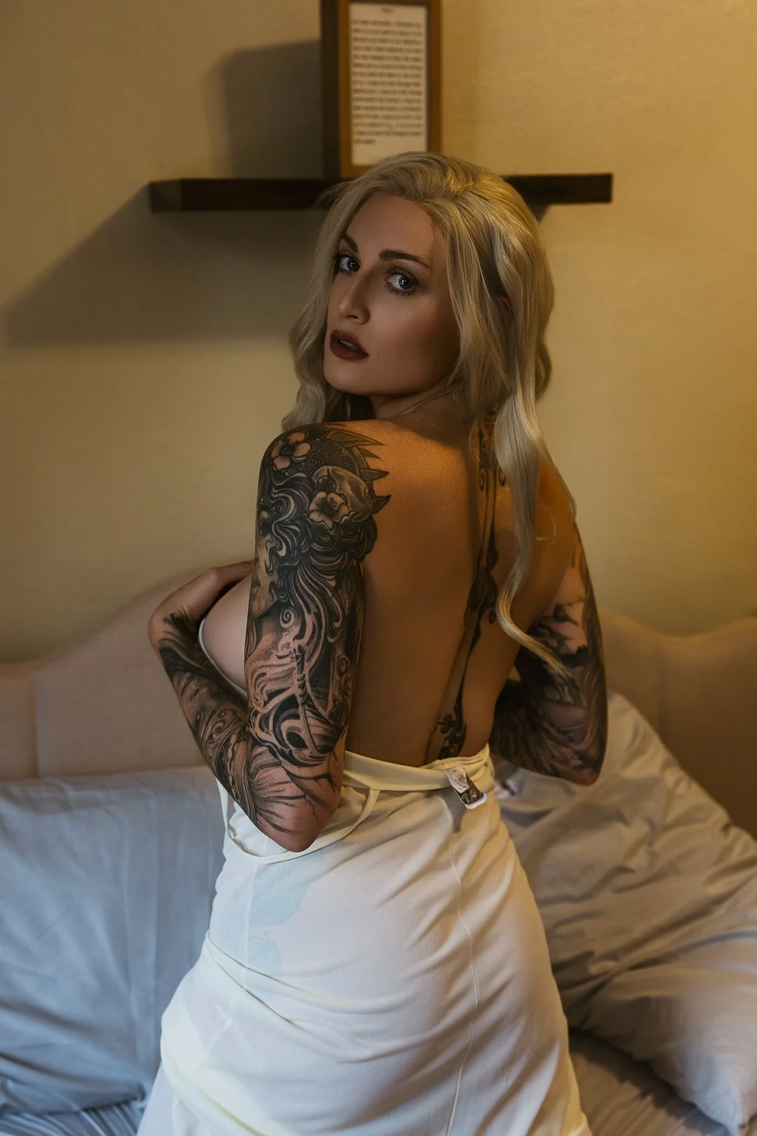 A woman with platinum blonde hair and tattoos on her arms and back posing in a hotel room, looking over her shoulder at the camera.