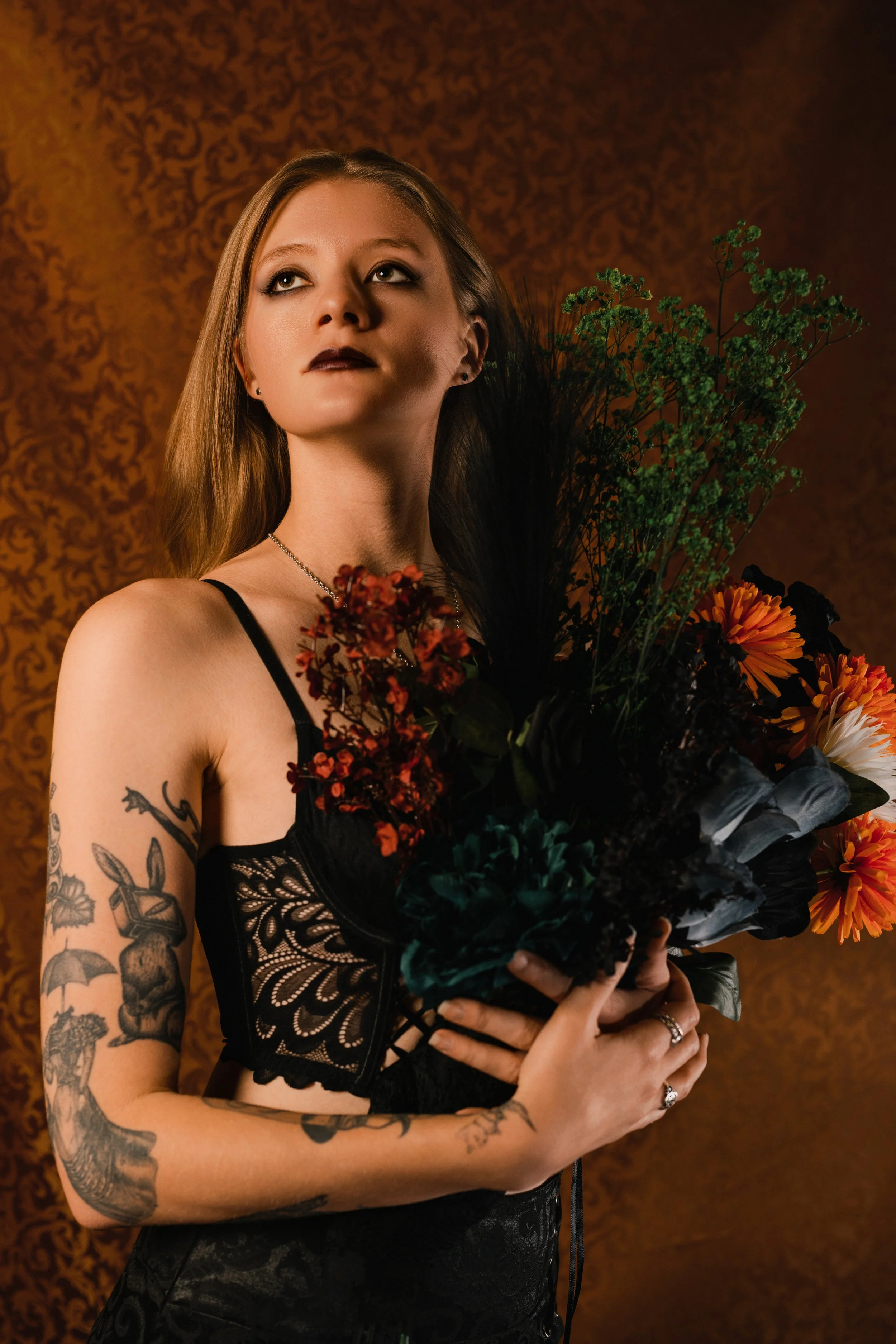 Young woman with long blonde hair, tattoos on her arms, wearing black lace lingerie, holding a bouquet of flowers, standing in front of a patterned orange-brown background.