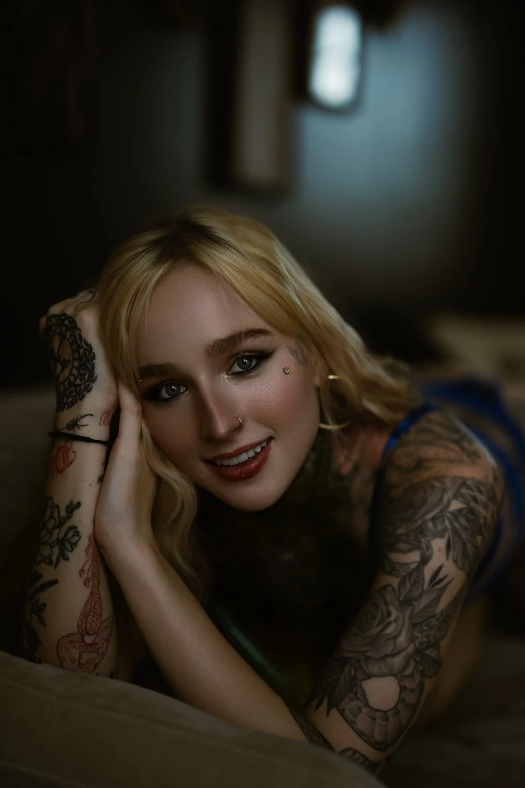 A young woman with blonde hair and tattoos is lying on her side, resting her head on one hand, and smiling at the camera.