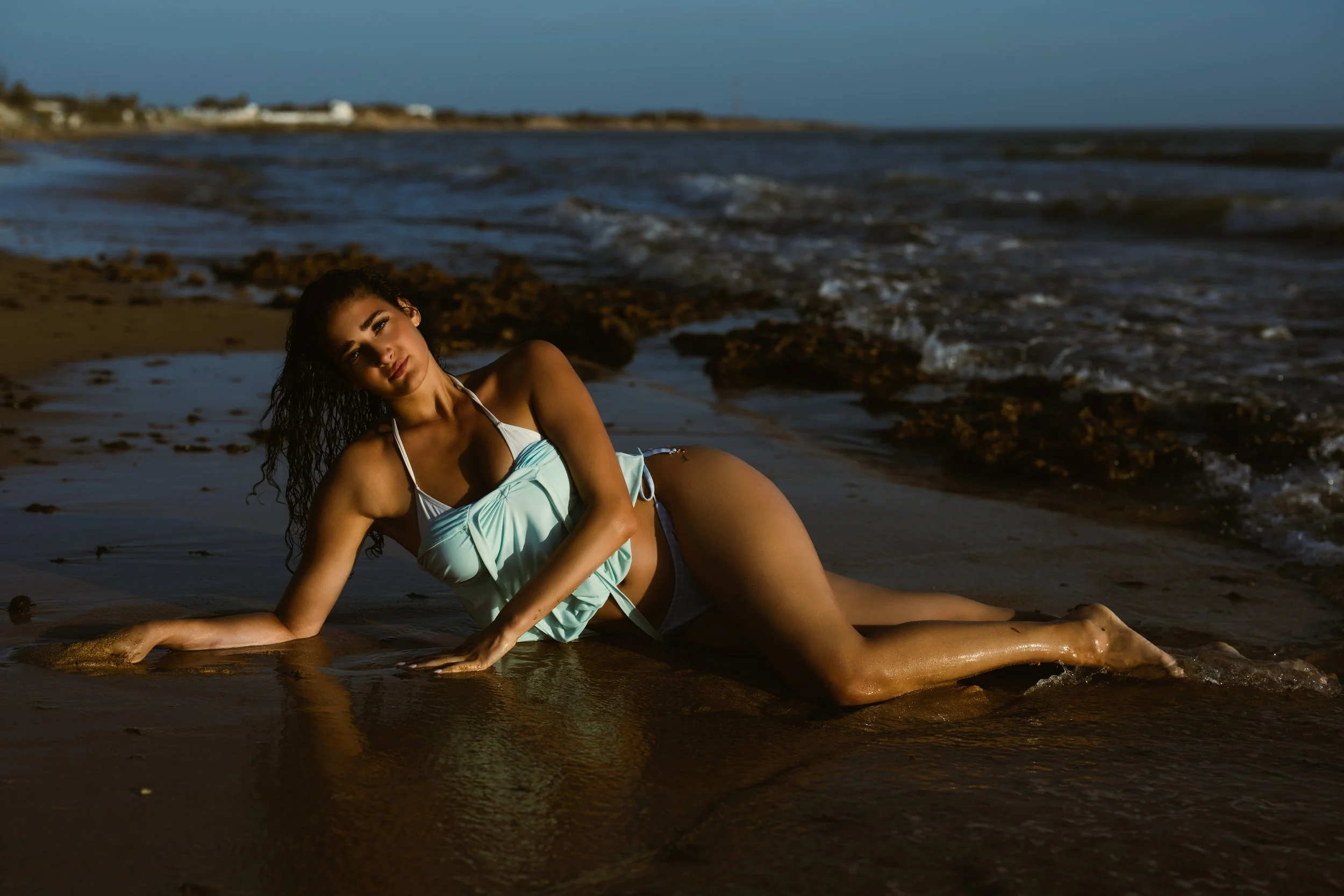 A woman with curly dark hair lies on her side on a sandy beach near the water, with rocks and the ocean in the background, during what appears to be sunset or dusk.