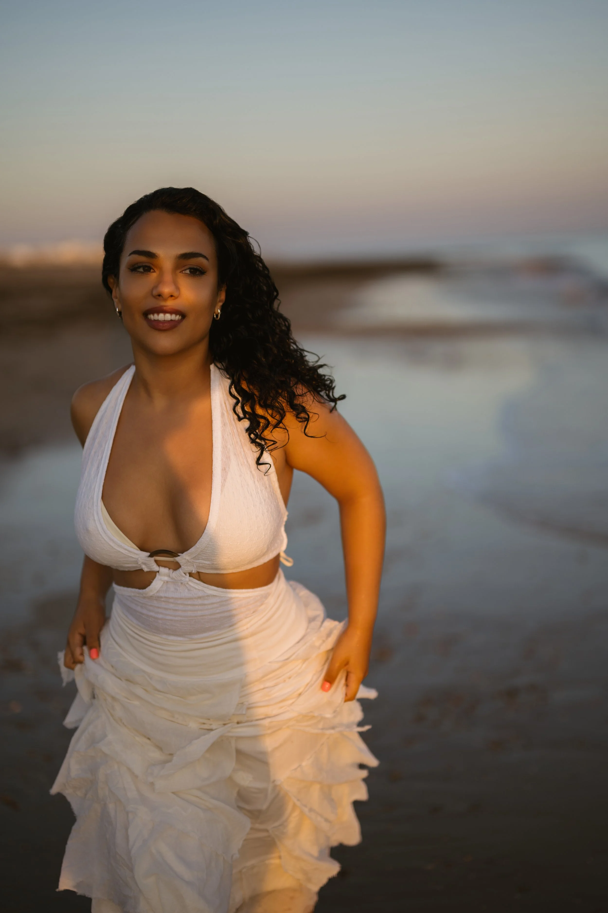 Young woman with curly dark hair and hoop earrings smiling at the camera on a beach during sunset, wearing a white crop top and flowing skirt.