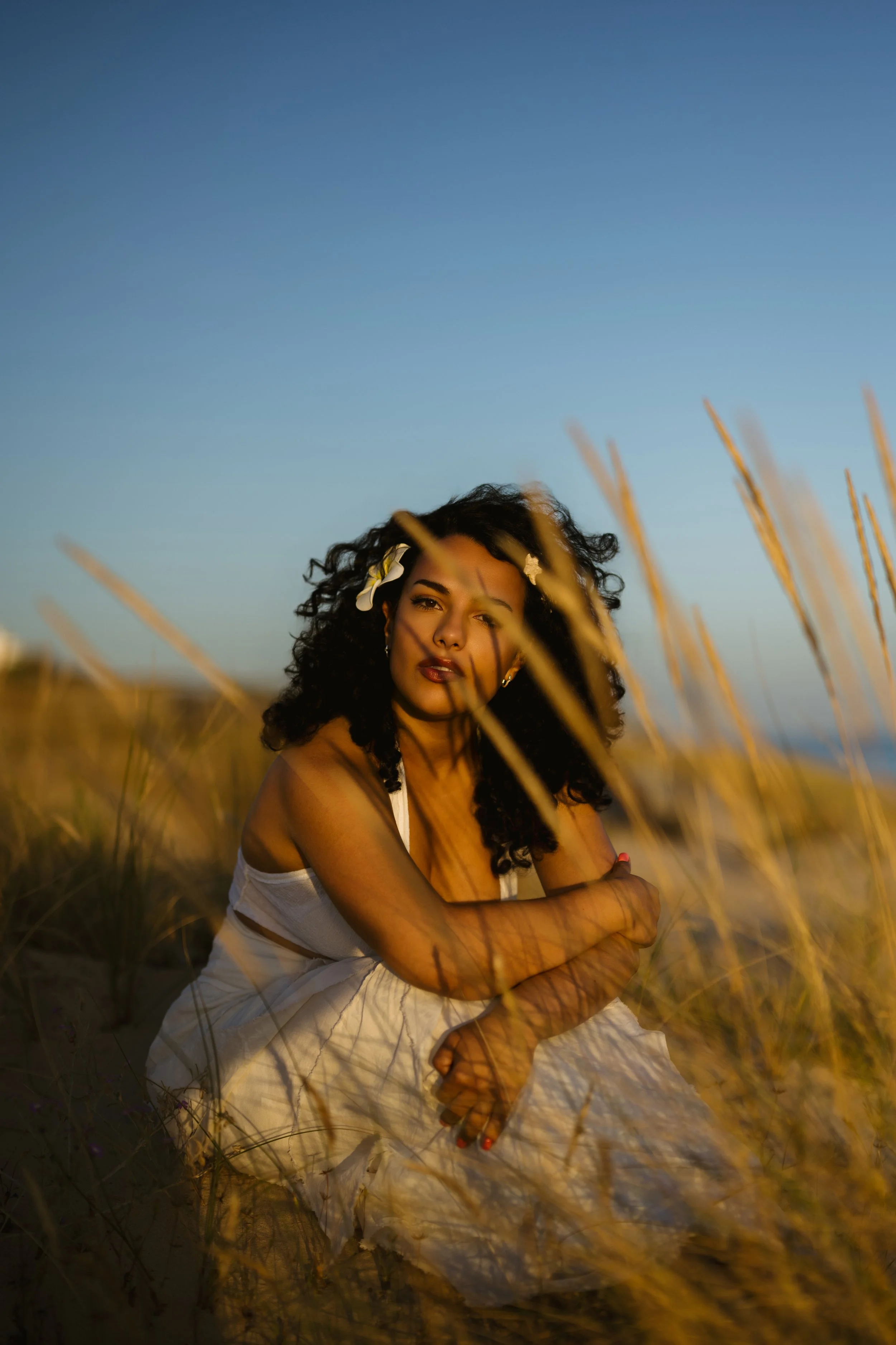 A woman with curly hair and star-shaped hair clips sitting in a field of tall grass during sunset, with a clear blue sky in the background.