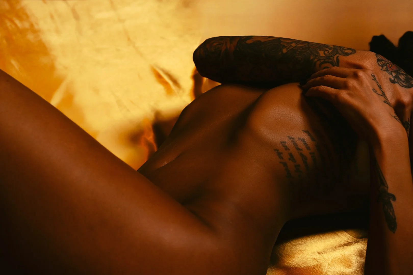 Close-up of a person with tattoos lying on a satin bed, with warm lighting creating a soft atmosphere.