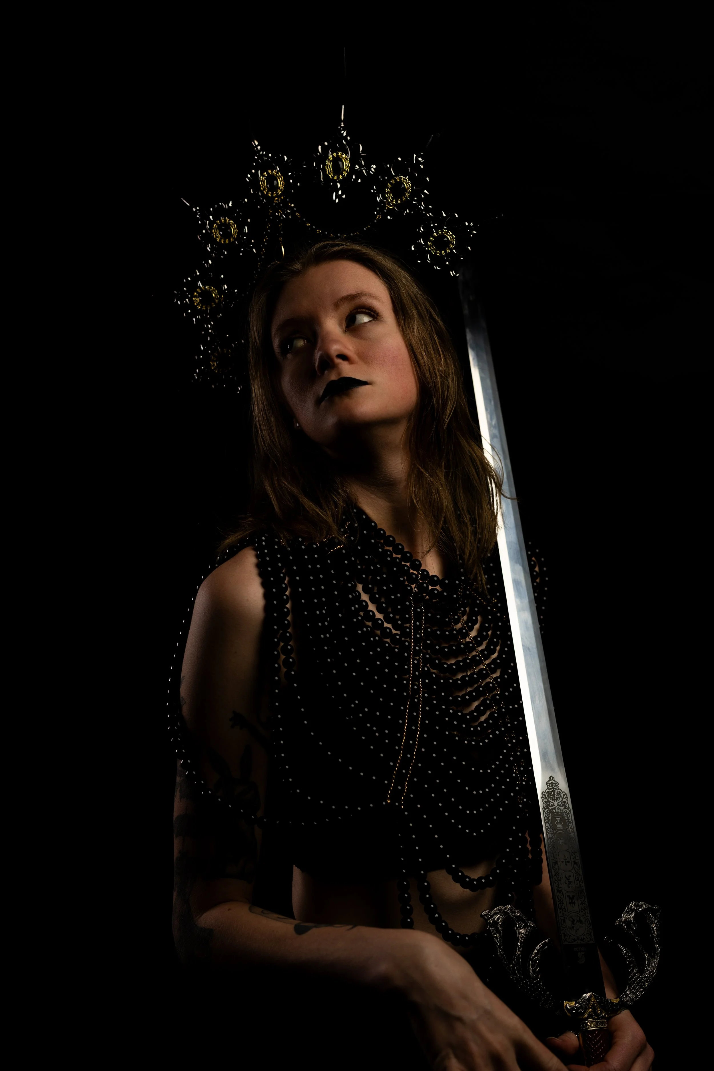 A young woman with long brown hair and black lipstick posing in a dark setting, wearing layered black pearl necklaces and a black polka dot sleeveless top, holding a katana sword with an intricate guard.