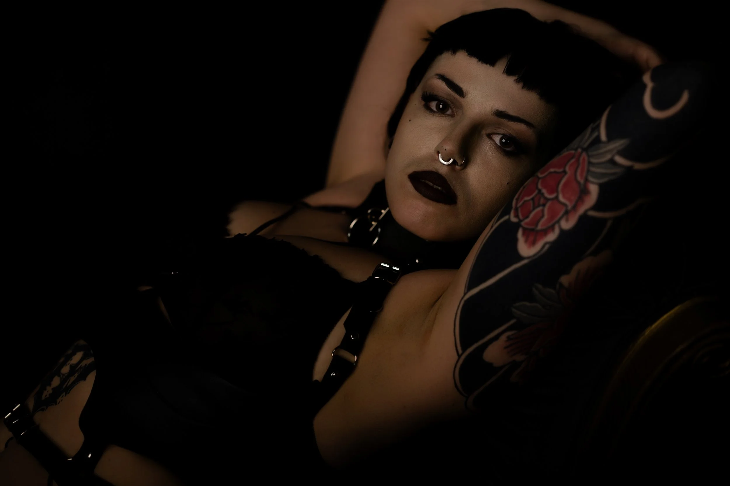 A person with short black hair, dark makeup, and facial piercings lying on a bed with black sheets, decorated with floral tattoos on their arm. The person has dark lipstick and is wearing a black choker.