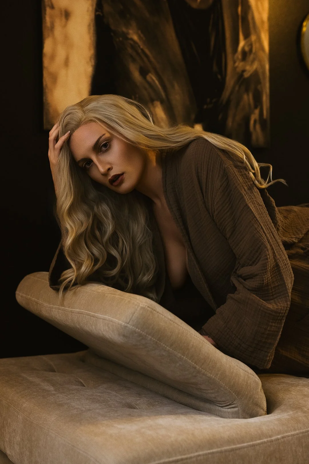 A woman with long, wavy blonde hair resting her head on a beige pillow, looking at the camera with a serious expression, in a dimly lit room with dark walls and abstract artwork in the background.