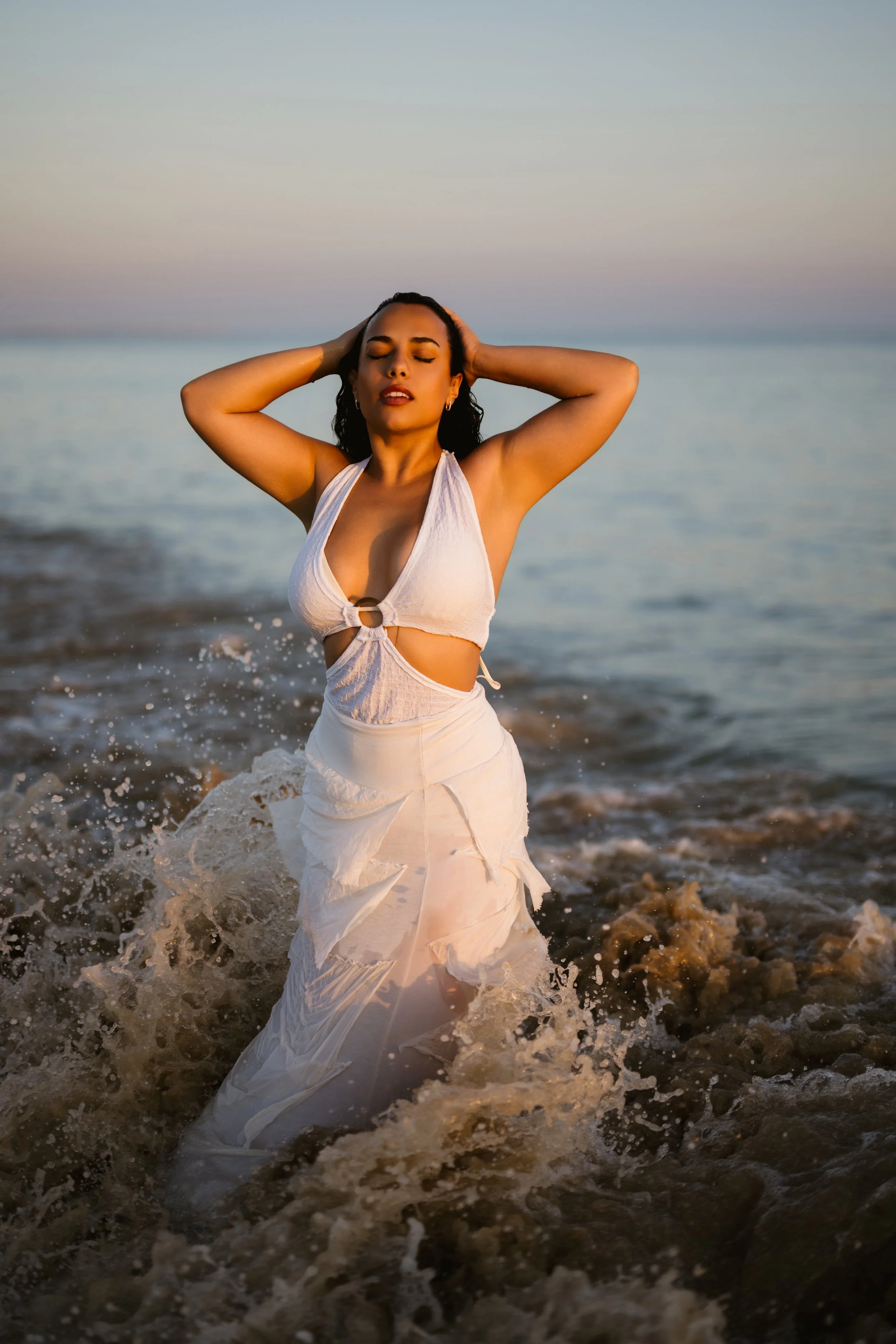 A woman in a white dress with a cut-out design stands in the water at the beach during sunset, with her eyes closed and hands on her head.