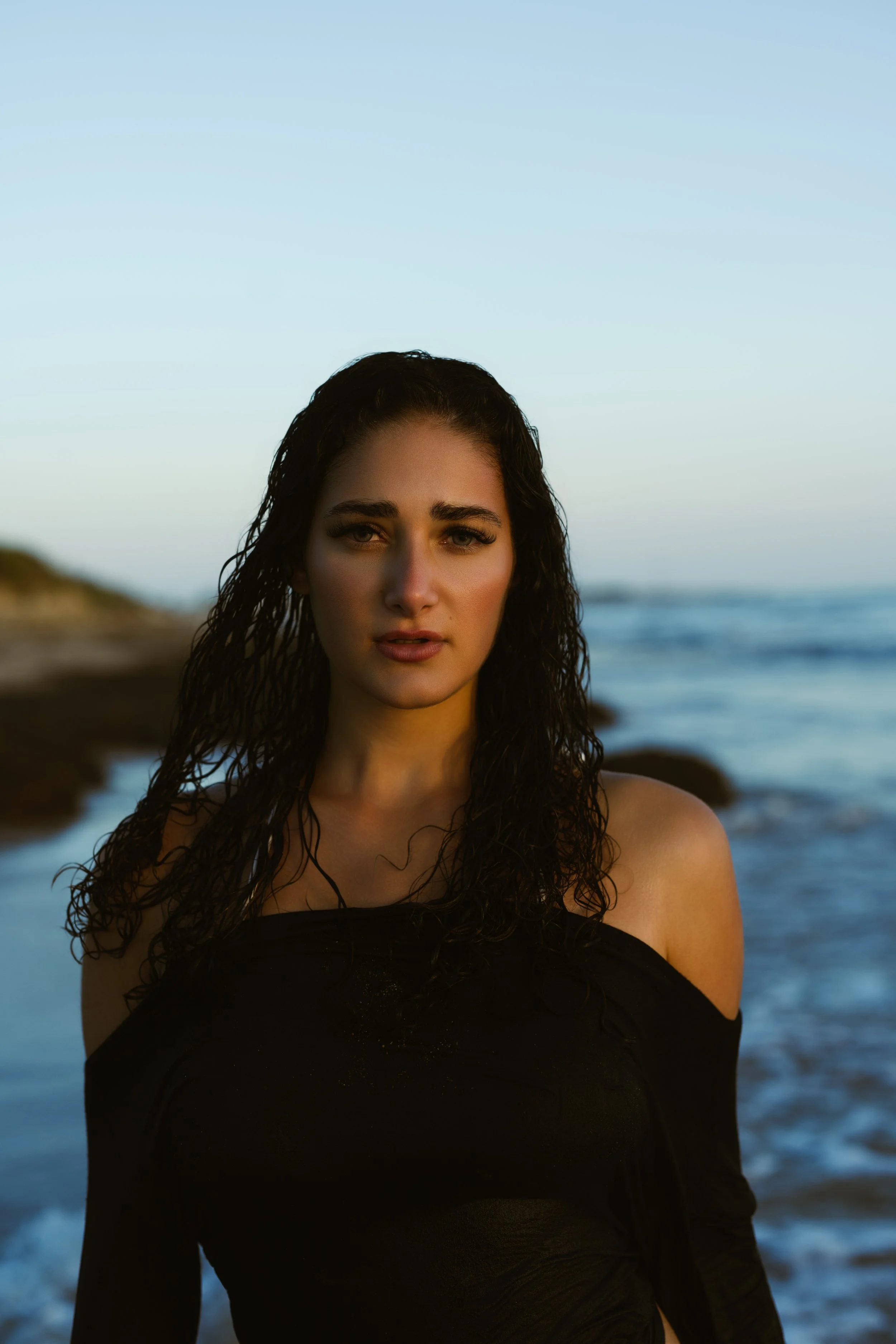 A woman with wet dark hair wearing a black top stands on a beach with the ocean and rocks in the background during sunset or sunrise.