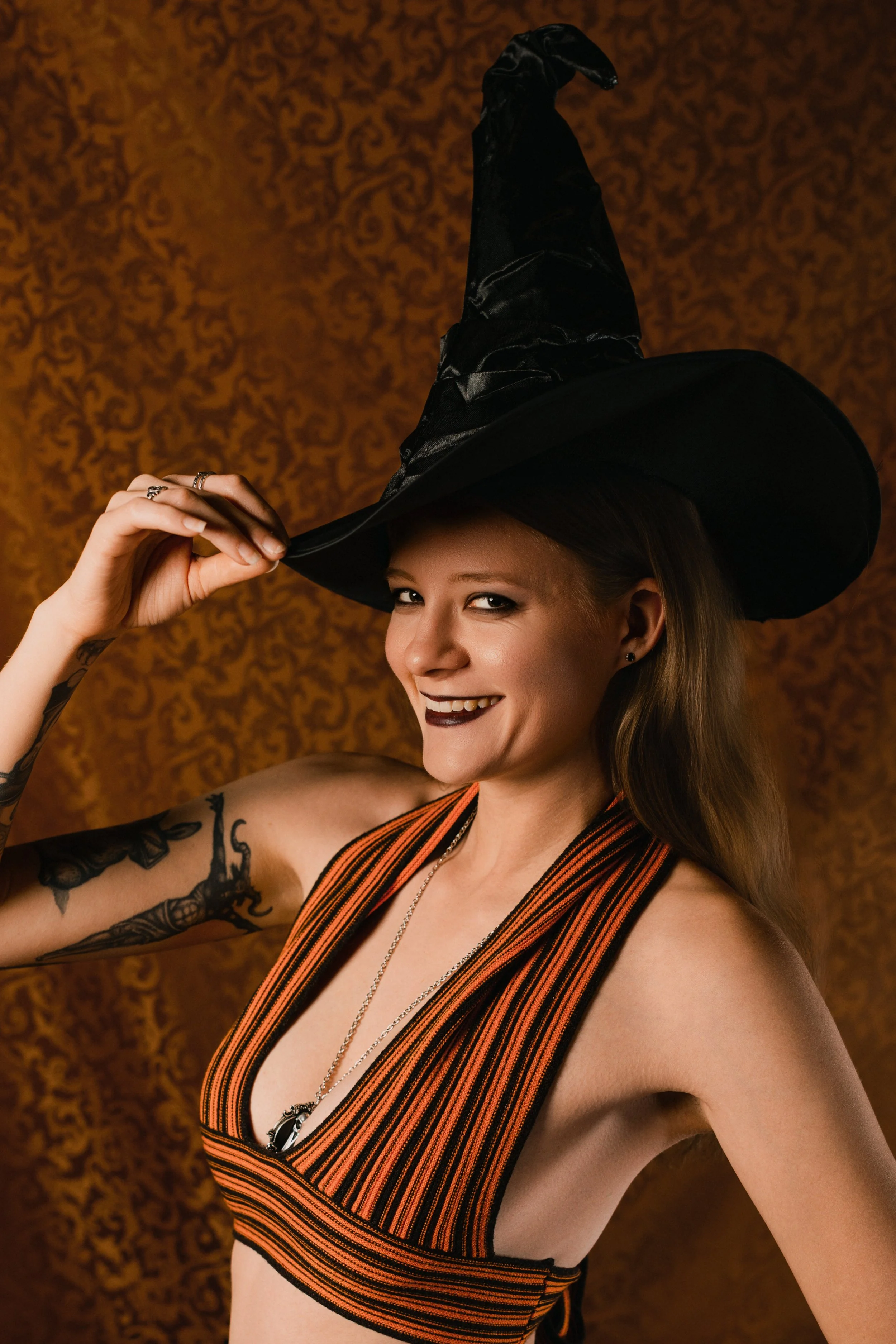 A woman in a black and orange striped top wearing a black witch hat, with tattoos on her arm, smiling against a brown patterned background.