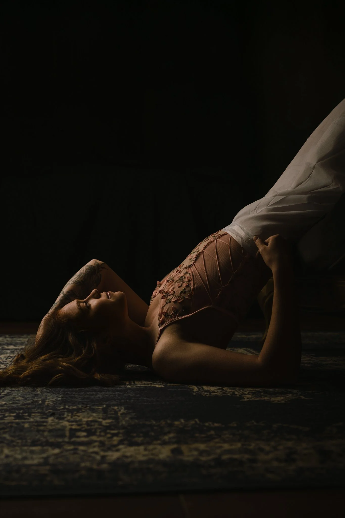 A woman with red hair lying on the floor, holding her foot, wearing a pink floral dress, and fishnet stockings, with tattoos on her arm, in a dimly lit room.