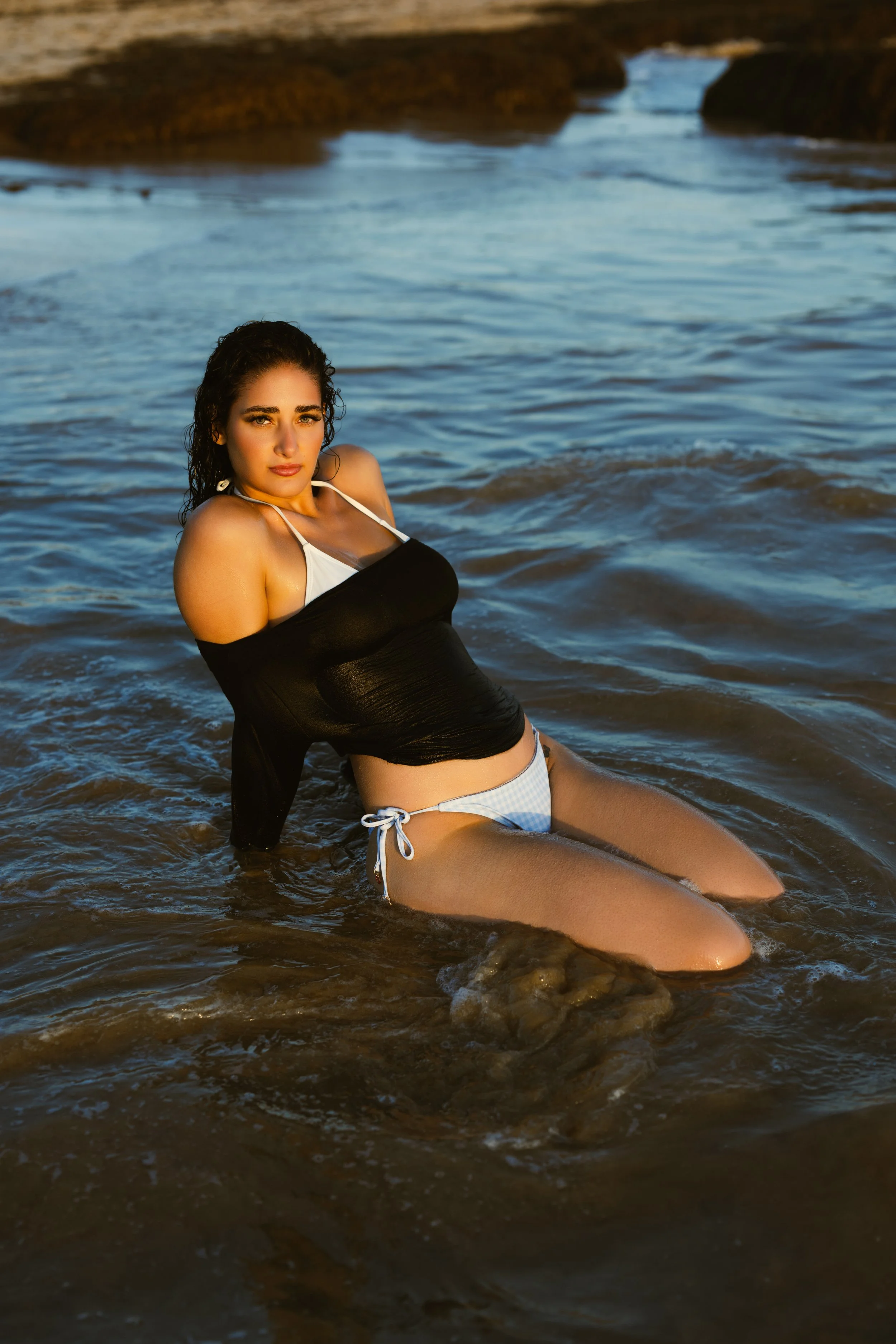 A young woman with dark curly hair and light skin sitting in shallow water at the beach during sunset, wearing a black and white swimsuit.