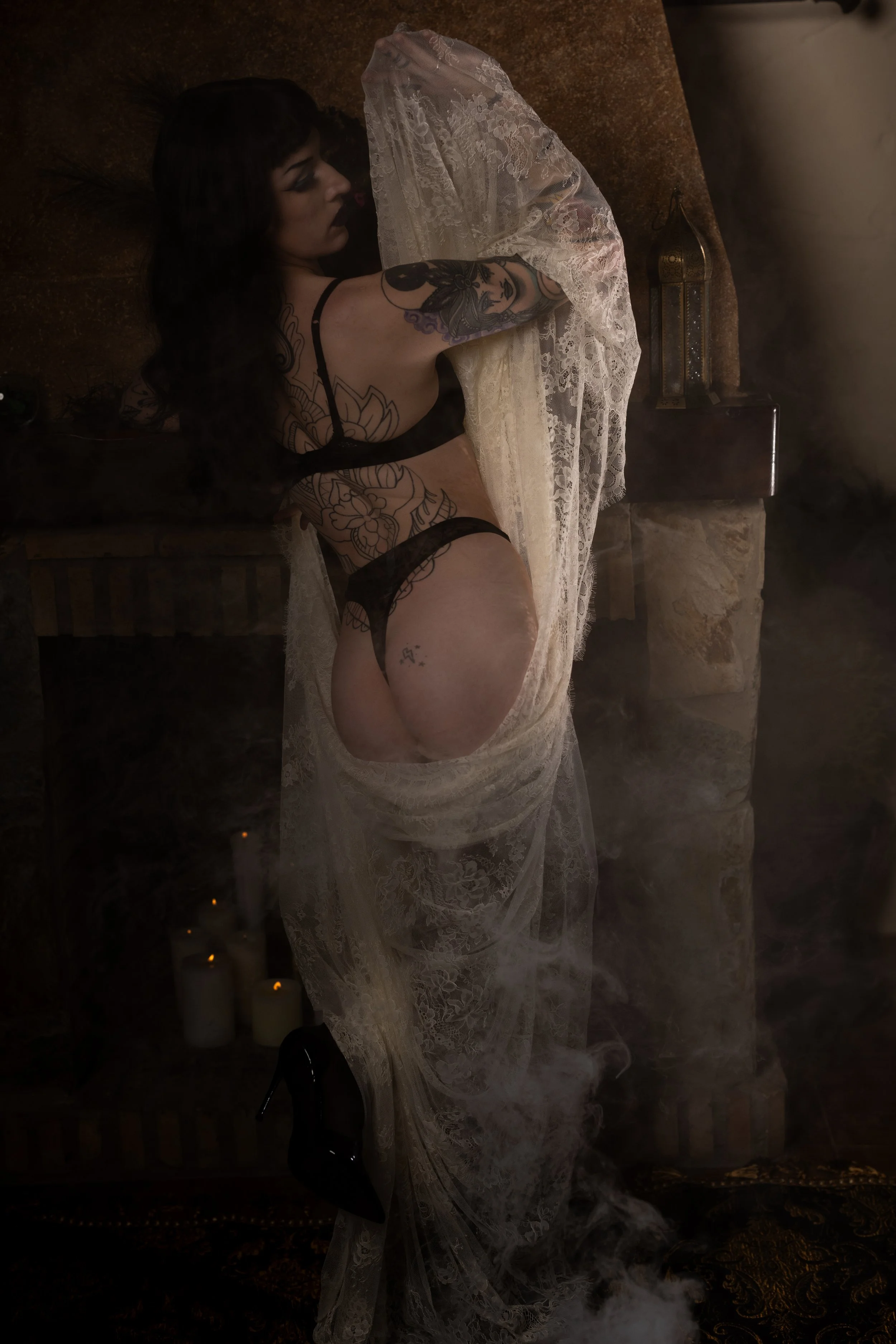A woman with tattoos and dark hair lying on a brown chaise lounge, partially covered by a white lace fabric, with lit candles on the floor and a rusty lantern in the background, surrounded by smoke or fog.