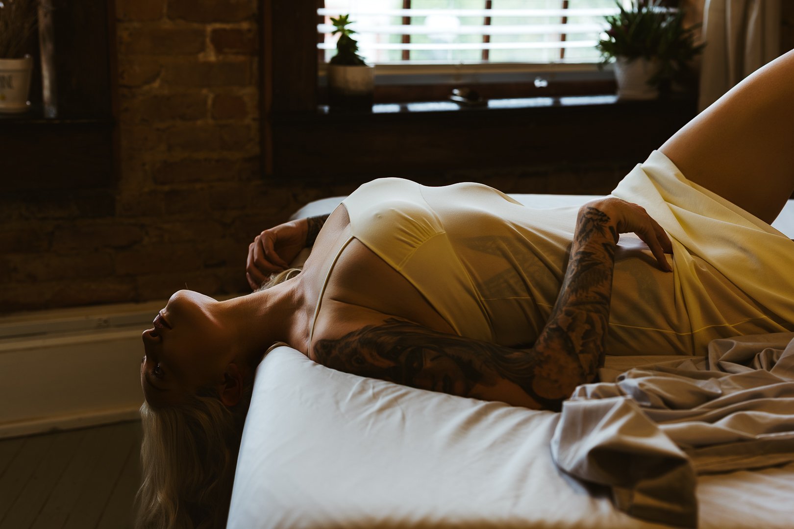 A woman with tattoos lying on her back on a bed, wearing a satin yellow slip, in a dimly lit room with a brick wall and window with blinds in the background.
