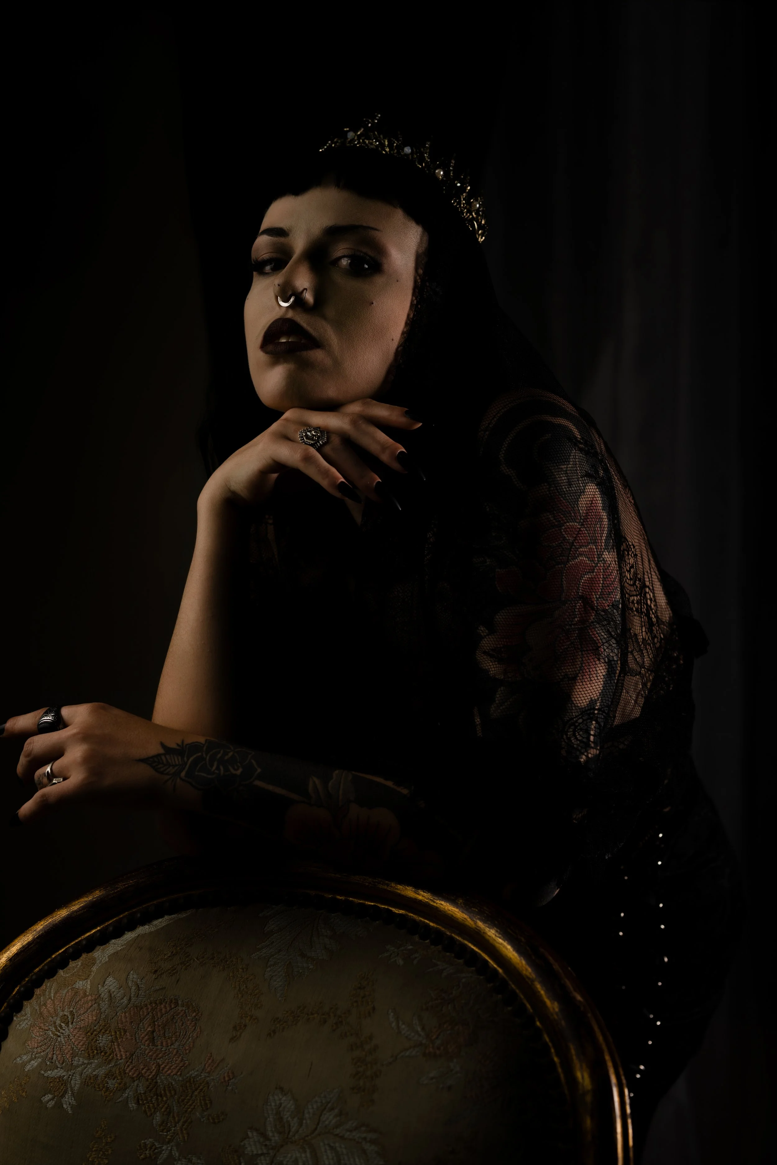 A woman with dark hair and makeup, wearing a tiara, seated at a table with a decorative cloth, in a dark setting.