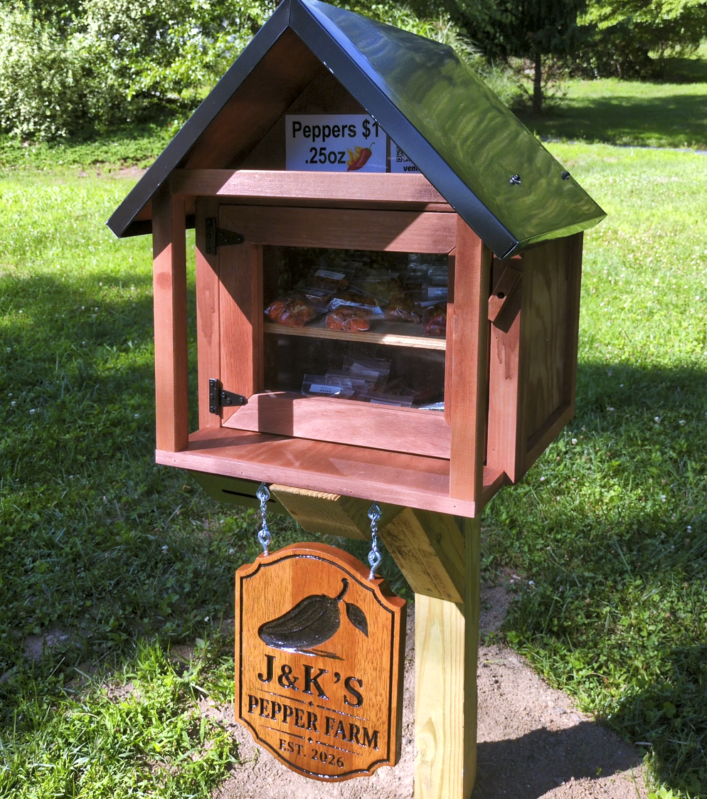 A wooden pepper stand shaped like a small house with a metal roof, located outdoors on a grassy area. The stand has a glass door displaying small bags of peppers and a sign above that reads "Peppers $1 .25oz". A decorative wooden sign hanging below says "J&K's Pepper Farm Est. 2026" with an image of a black pepper.