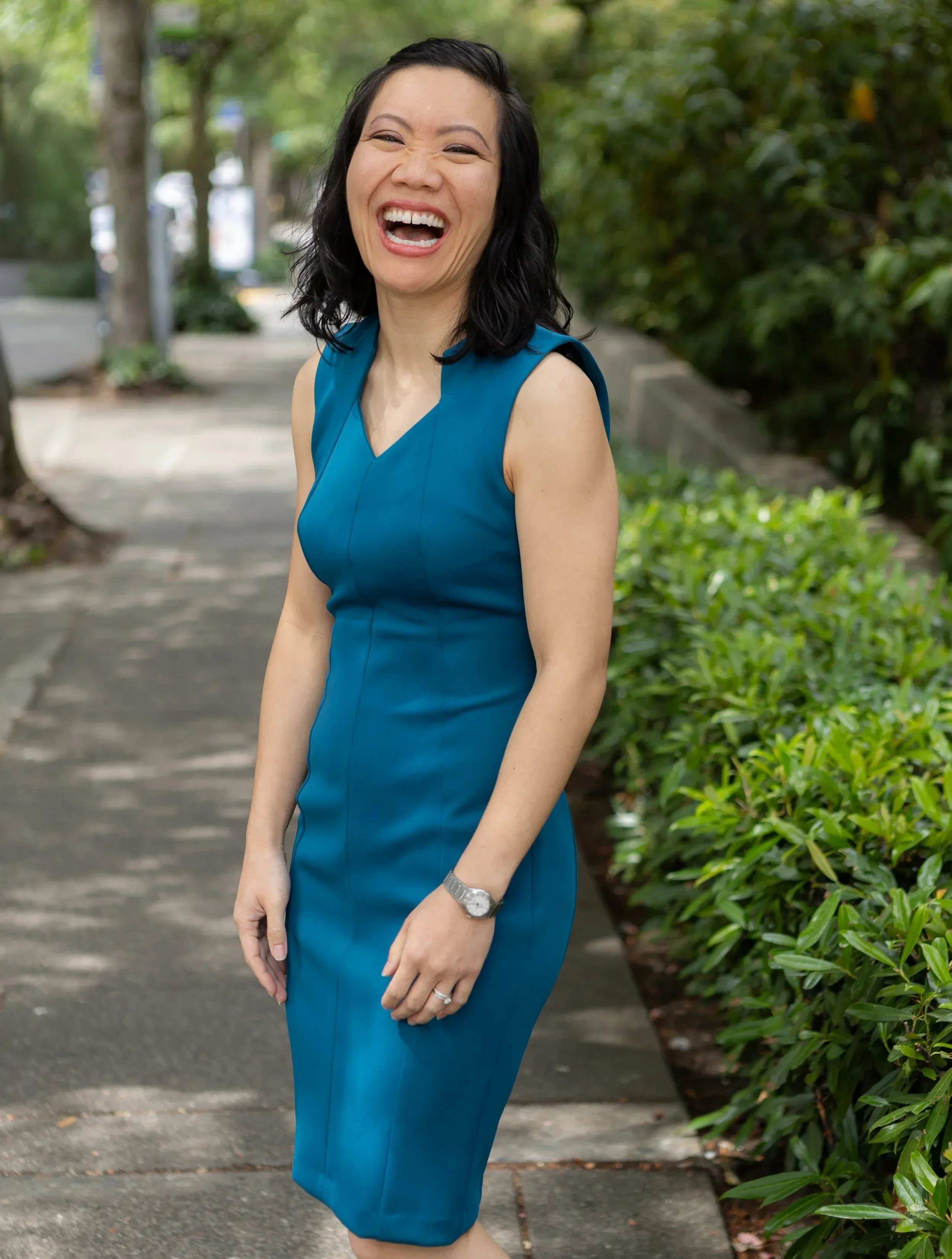 Dr. Judy Vu in a blue dress smiling and laughing outdoors on a sidewalk with green bushes and trees in the background.