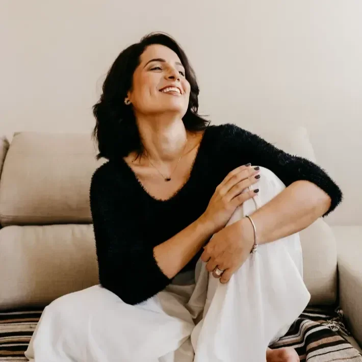 A woman with dark hair smiling and sitting on a couch, wearing a black top and white pants.