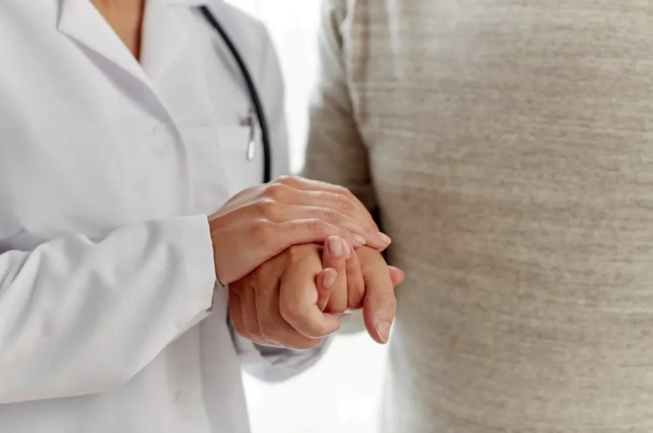 A healthcare professional, dressed in a white coat, holding a patient's hand in a comforting gesture.