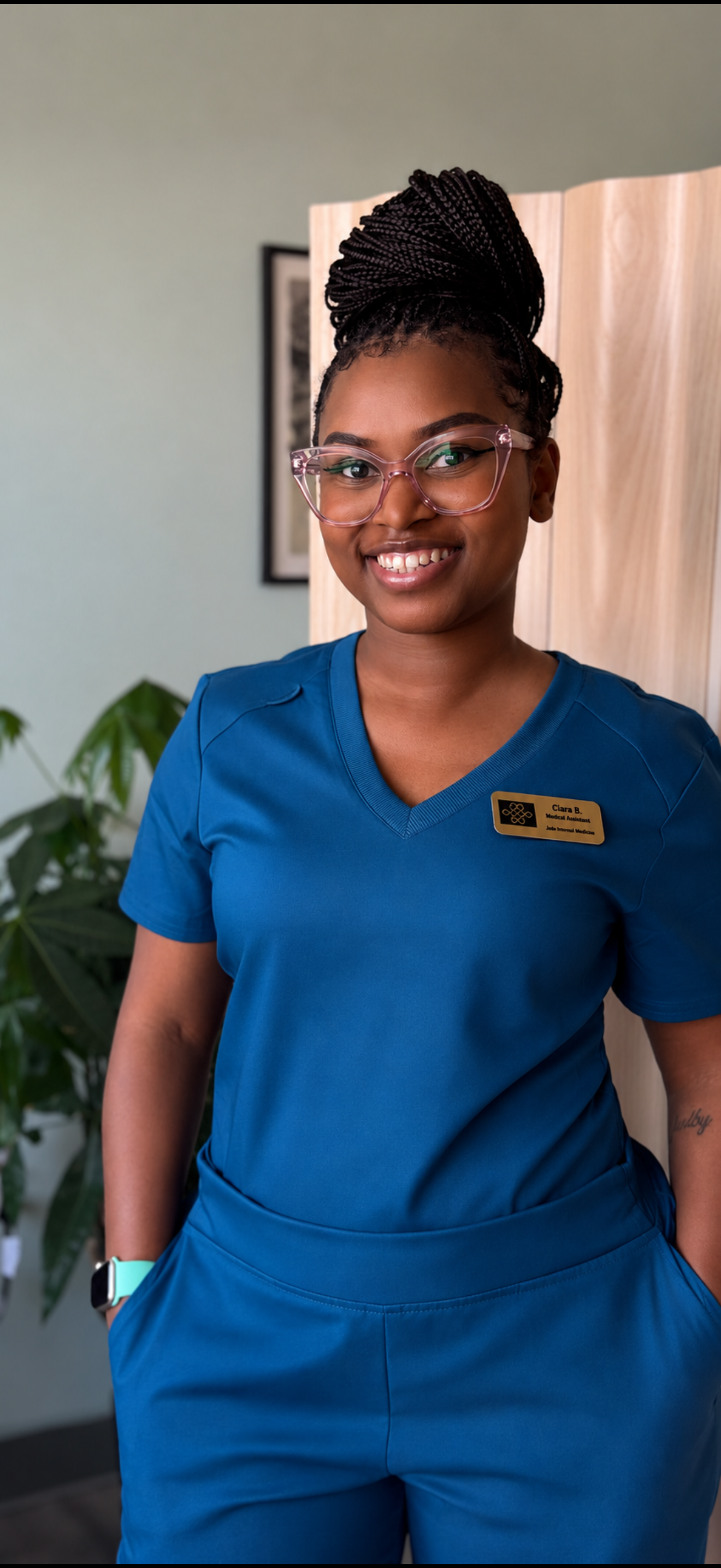 Ciara Barney wearing blue medical scrubs and a name tag, standing in front of a wooden divider and green plant.