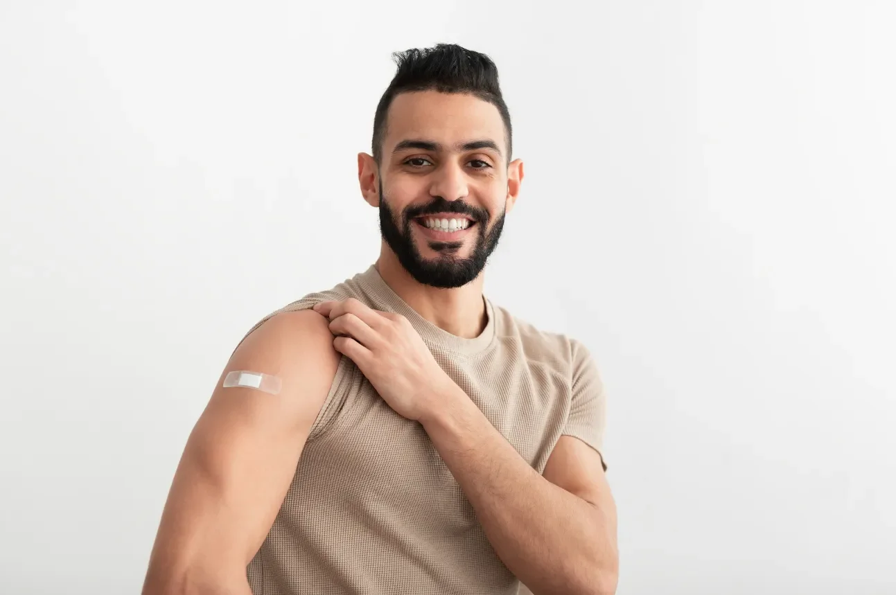 Man smiling and showing shoulder with a bandage in a beige sleeveless shirt, standing against a plain white background.