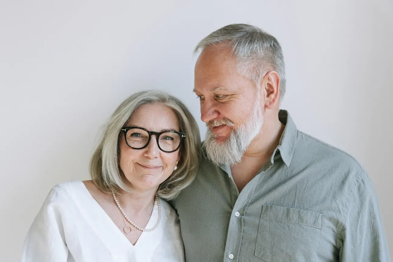 An older woman with blond hair, glasses, and a pearl necklace smiling, standing close to an older man with gray hair and a gray beard, who is looking at her with a gentle smile against a plain white background.