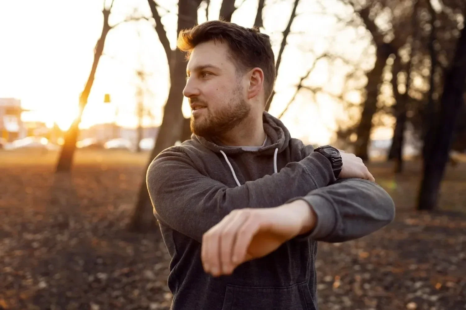 A man in a gray hoodie stretching his arms in a park during sunset, with trees and blurred buildings in the background.