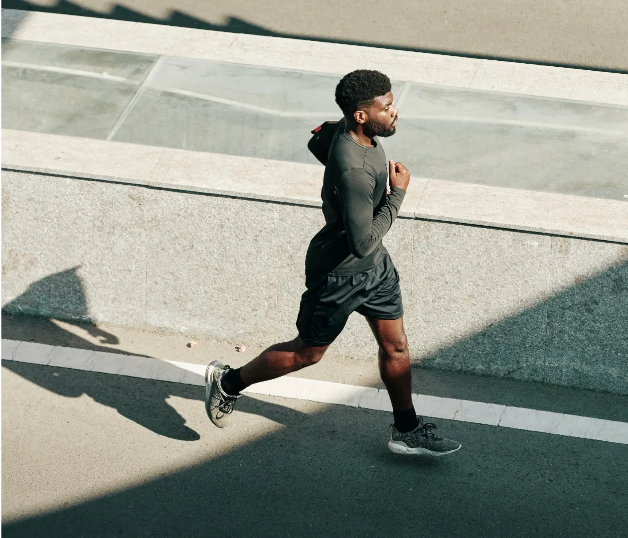 A young man running outdoors on a city street during daytime, wearing black athletic clothing and running shoes.