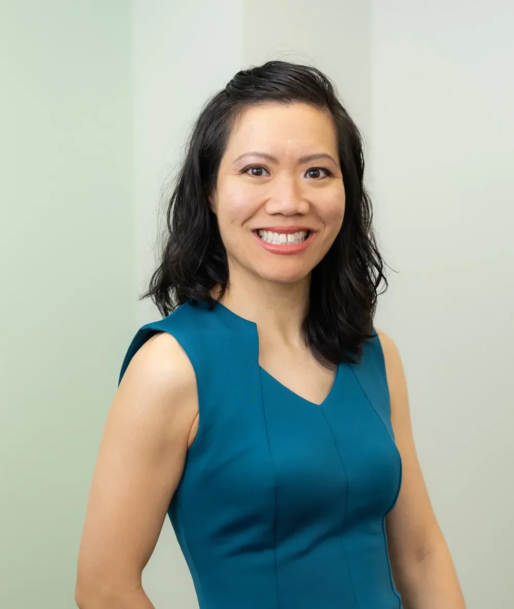 Dr. Judy Vu with shoulder-length black hair, smiling, wearing a sleeveless teal dress, standing against a light green background.