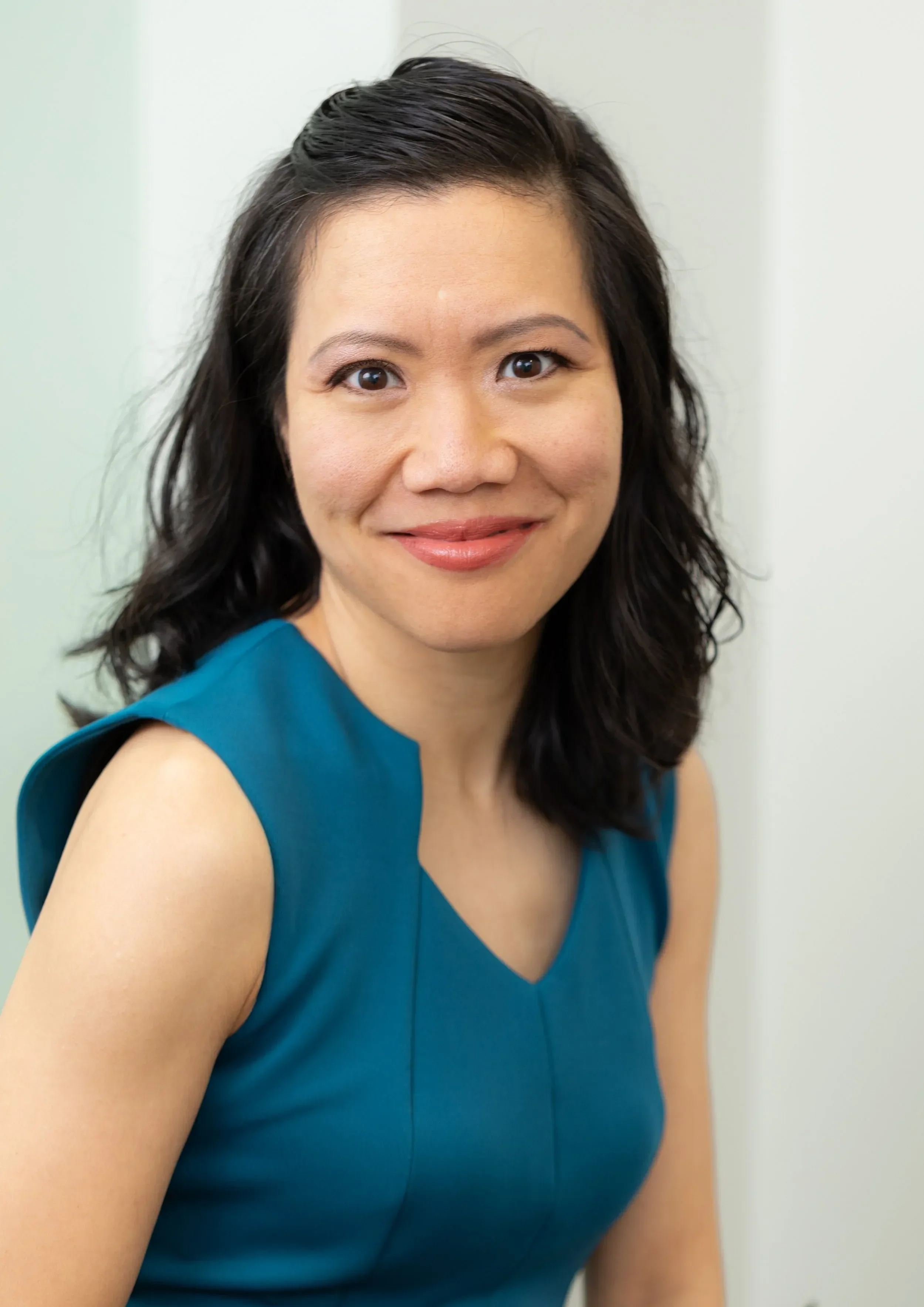 Close-up portrait of Dr. Judy Vu with dark wavy hair wearing a teal sleeveless top, standing against a light-colored wall.
