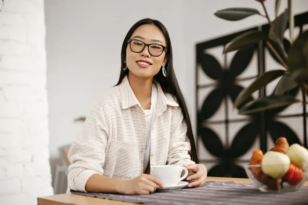 A woman with long dark hair, glasses, and earrings sitting at a table holding a white mug, smiling in a bright, modern interior with plants and white brick wall.