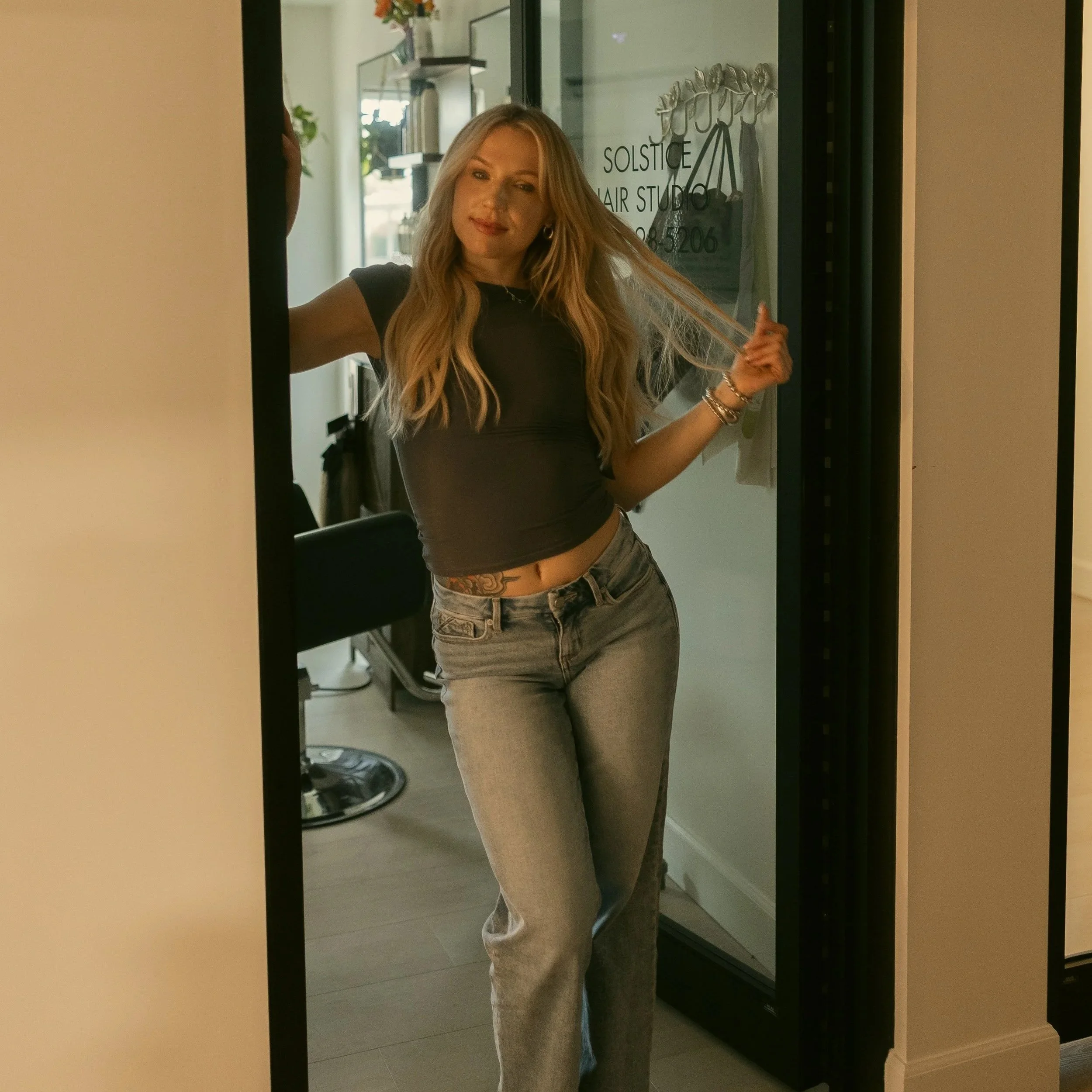 The owner of Solstice Hair Studio, Christian Taylor, with long blonde hair, wearing a black top and jeans, with a sign that reads 'Solstice Hair Studio' in the background.
