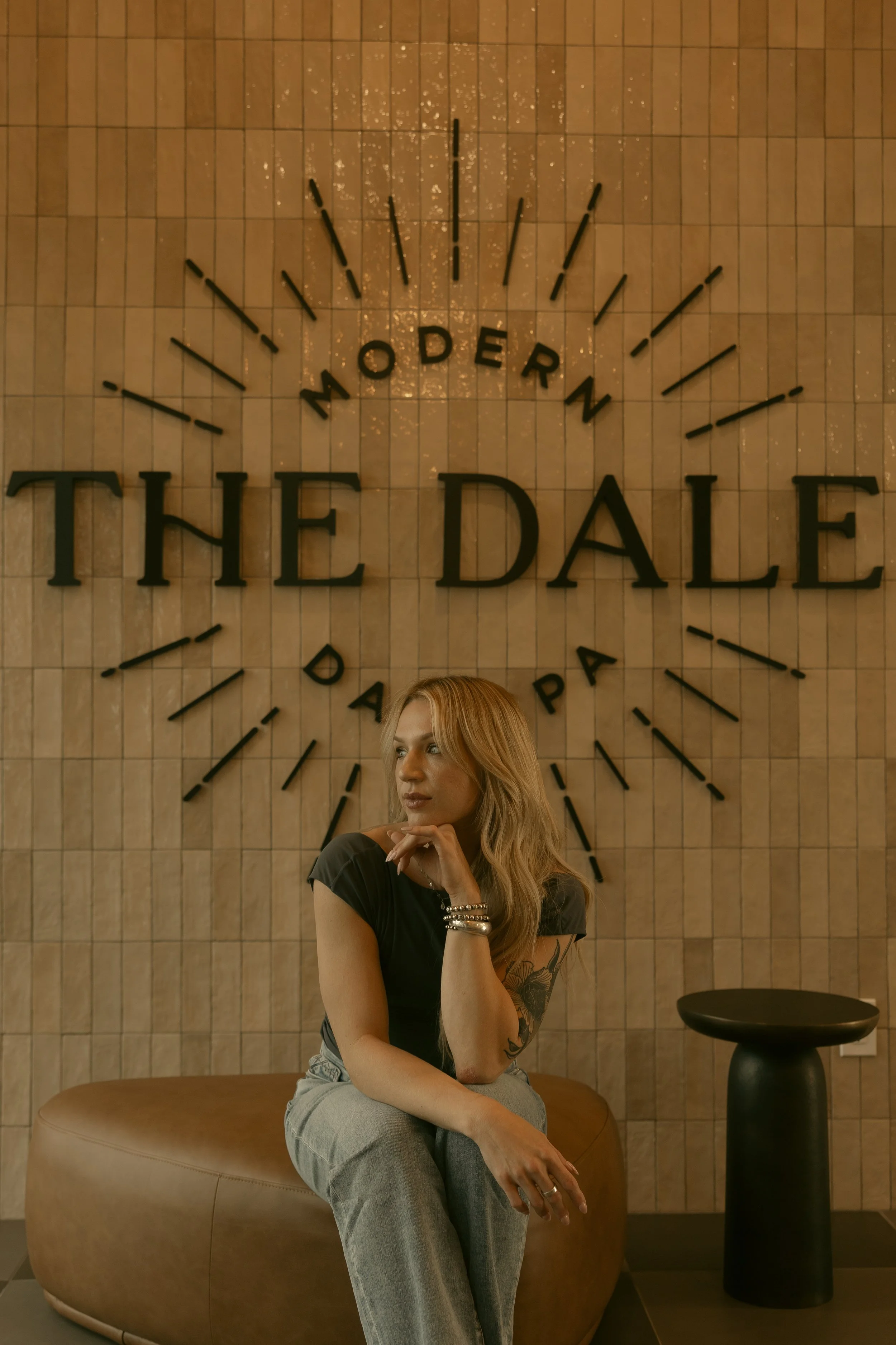 The owner of Solstice Hair Studio, Christian Taylor, sitting on a round, brown bench with her left hand resting near her chin, in front of a wall with a large clock that spells out 'THE DALE'.