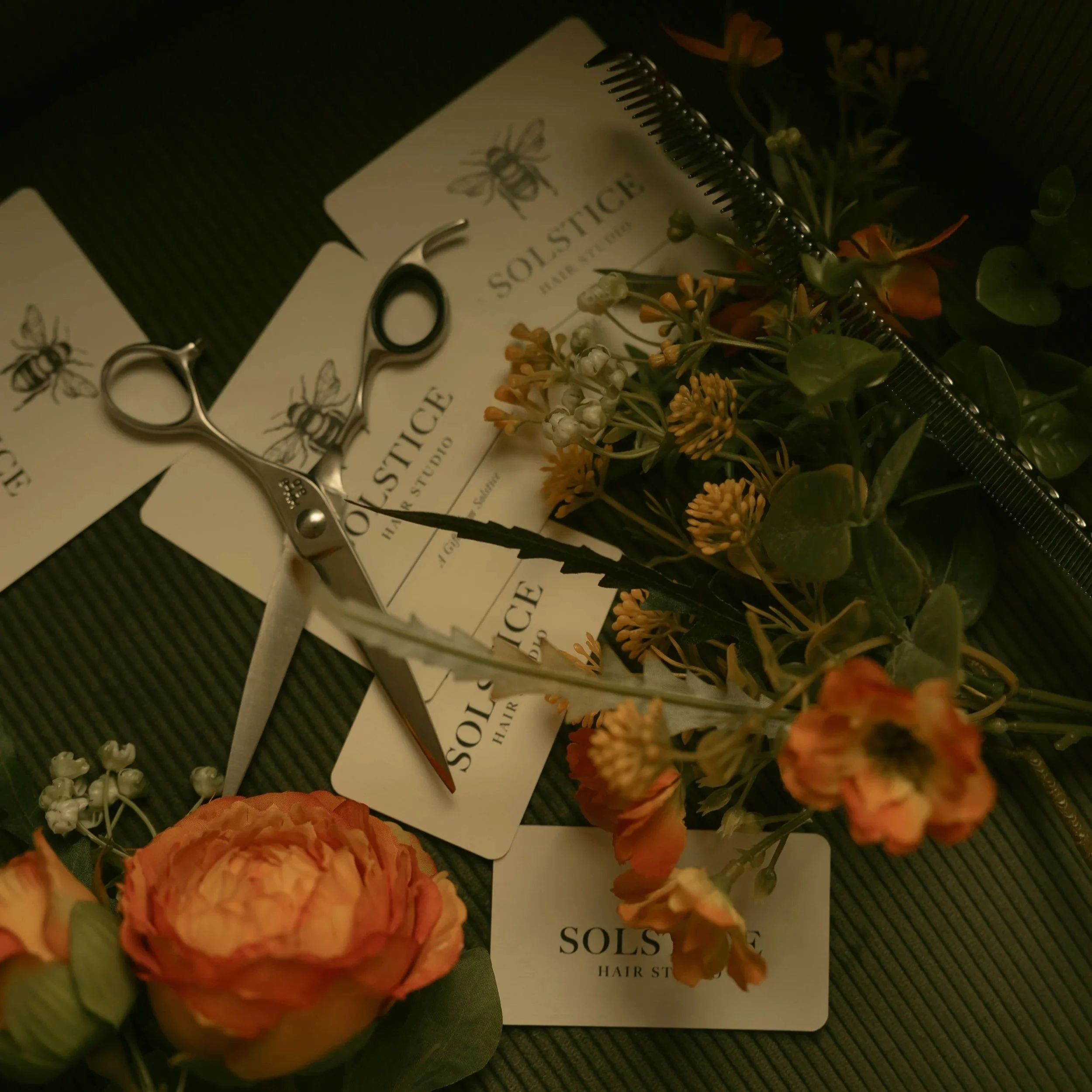 A close-up of flowers, a hair scissors, and Solstice Hair Studio business cards on a dark surface.